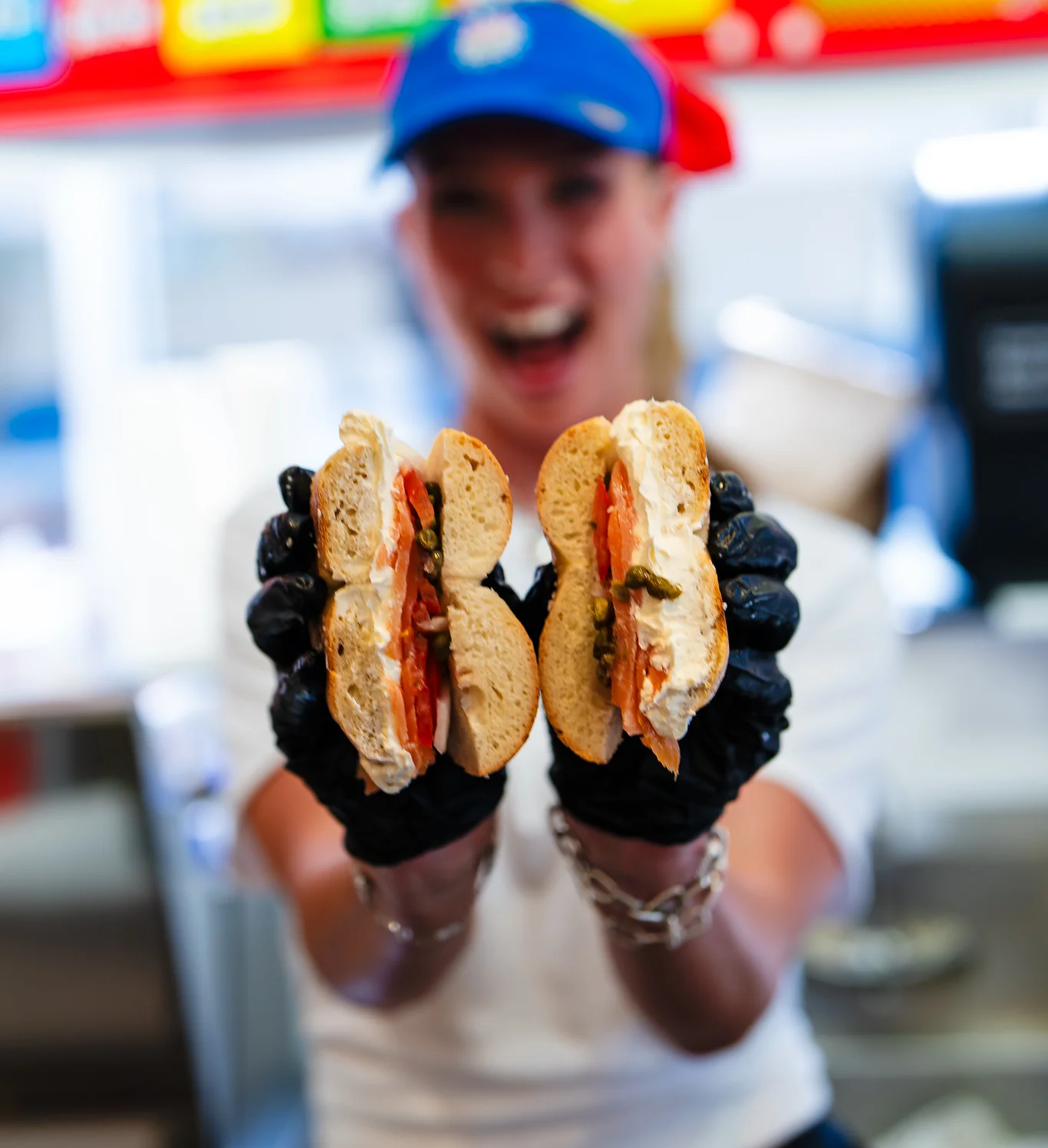 Employee holding two sandwiches with toppings, smiling in a restaurant setting.