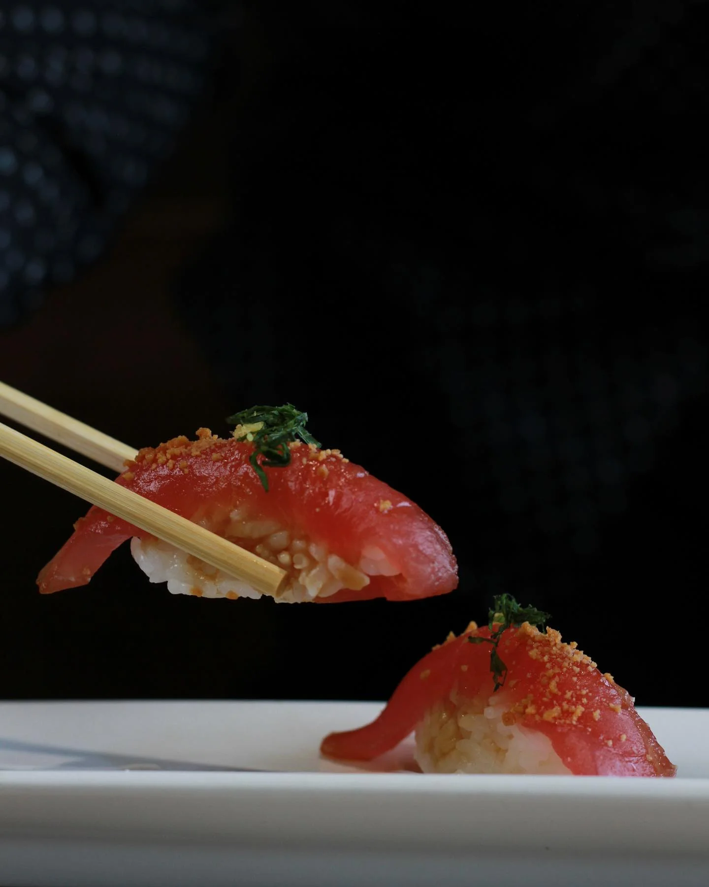 Tuna nigiri garnished with crunchy toppings and fresh herbs, being picked up with wooden chopsticks against a dark background.