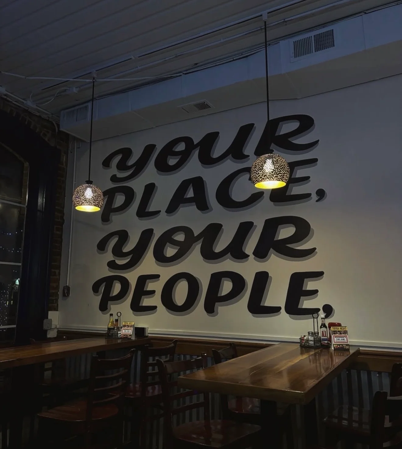 A cozy dining interior showcasing a wall with an inspiring quote and stylish pendant lighting. Simple wooden tables and chairs are set in the foreground.
