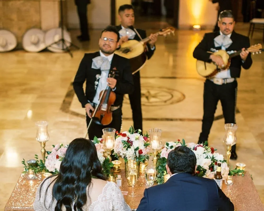 A mariachi band performing at an event with a beautifully decorated table in the foreground featuring flowers and candles.
