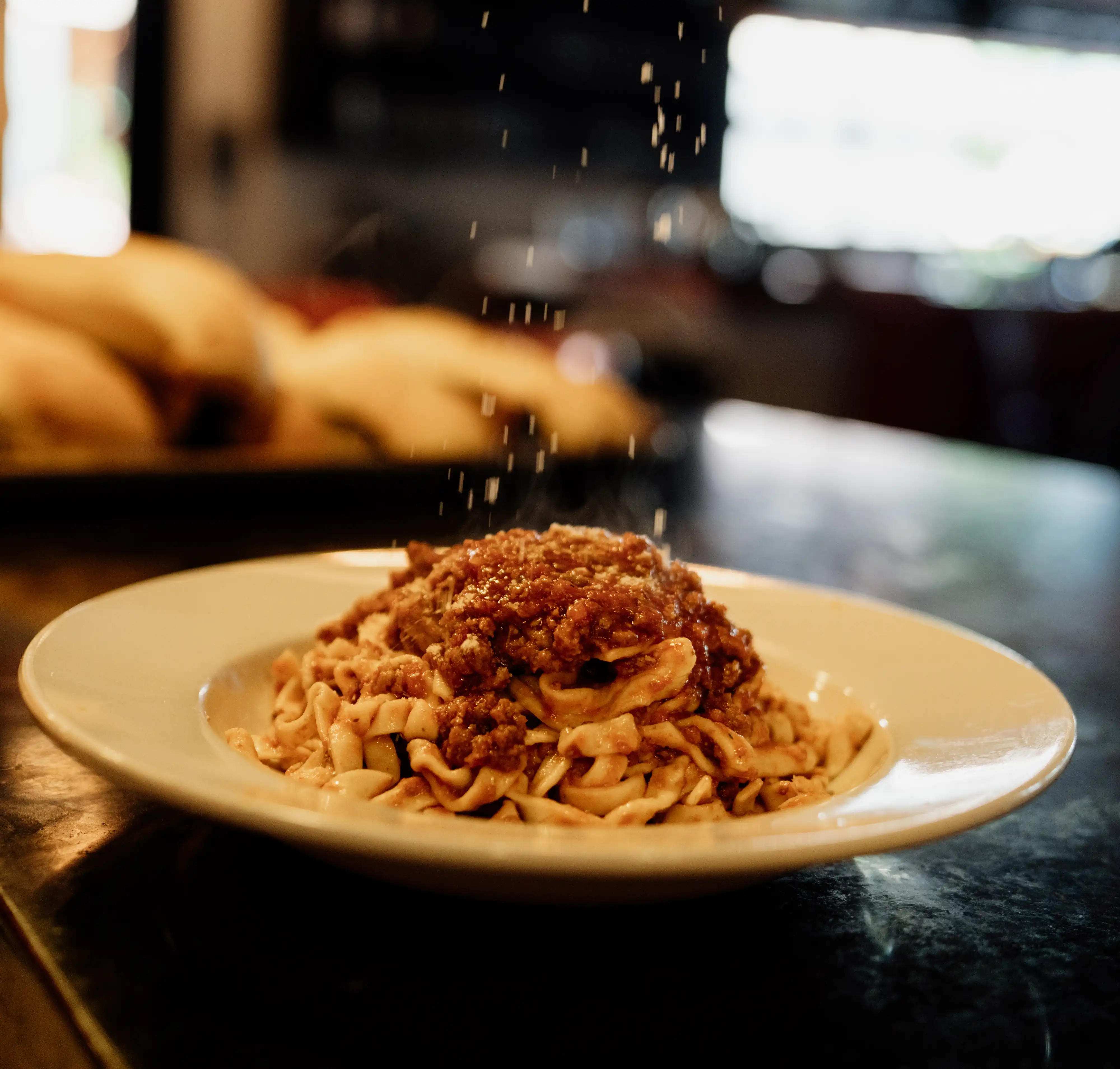 sprinkles of parmigiano cheese falling onto fresh bolognese pappardelle noodles on a black countertop