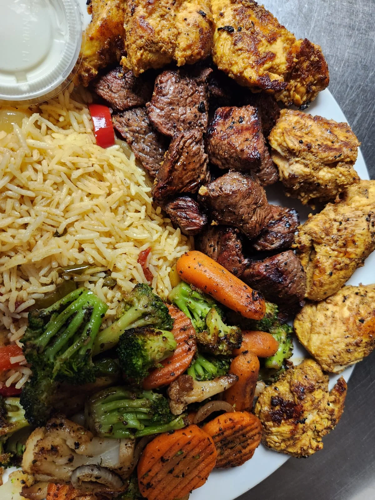 A hearty platter featuring grilled meats, rice, and assorted vegetables, served with a portion of sauce.