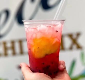 A colorful layered drink served in a clear cup, featuring shades of red and orange with ice cubes and a straw.