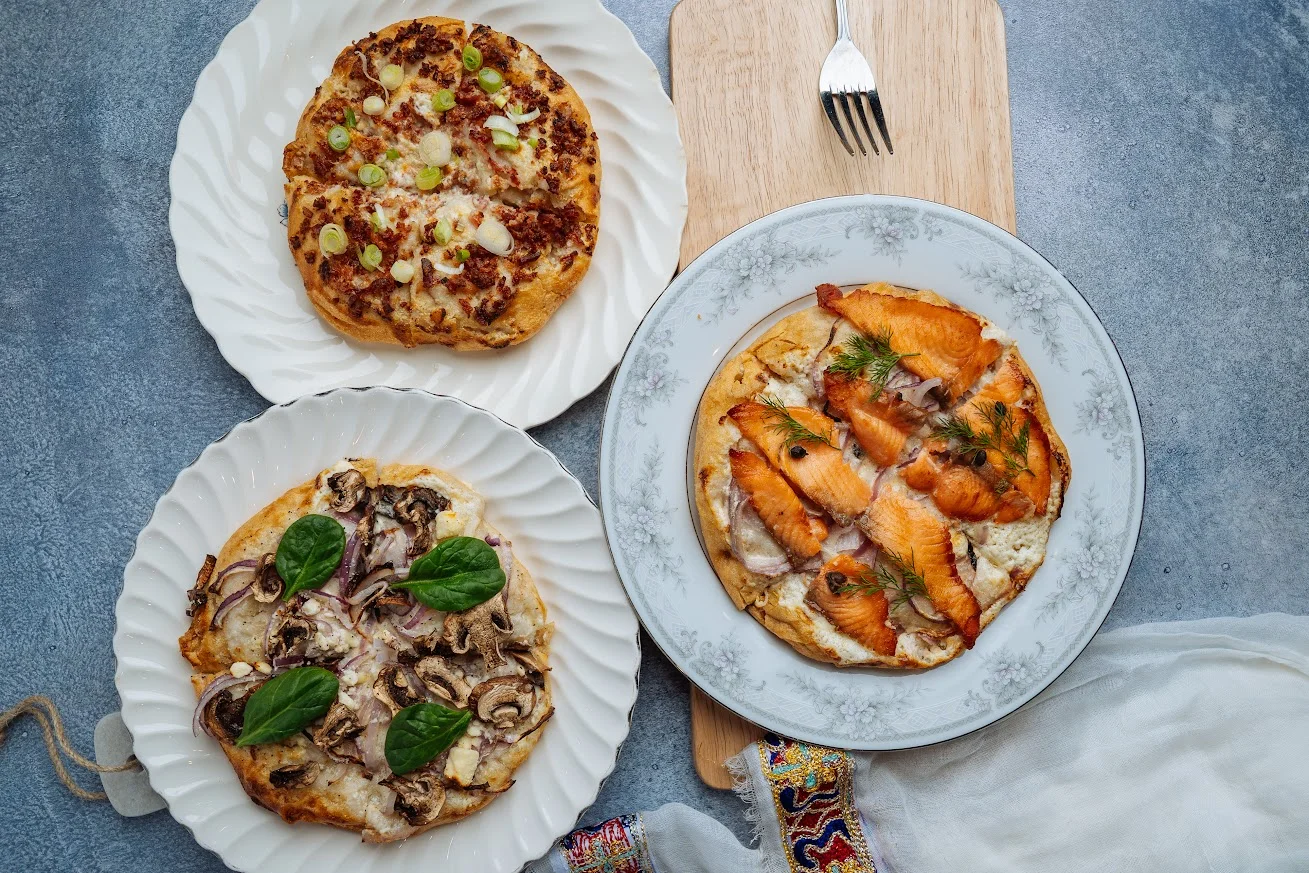 Three different types of flatbreads topped with various ingredients such as mushrooms, salmon, and green onions, presented on elegant plates.