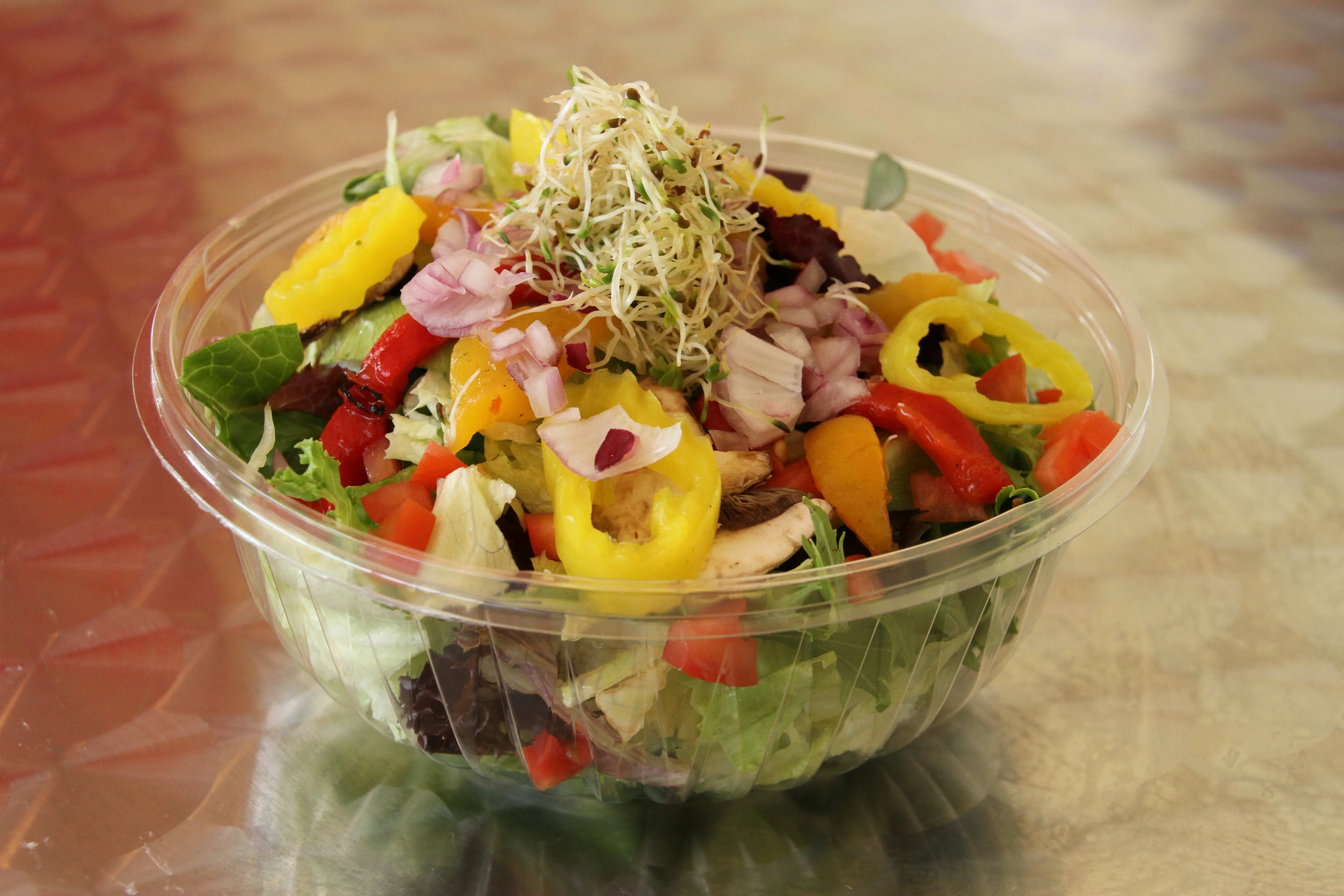 Fresh garden salad in a clear plastic bowl, topped with banana peppers, diced red onions, red bell peppers, shredded sprouts, and mixed greens.