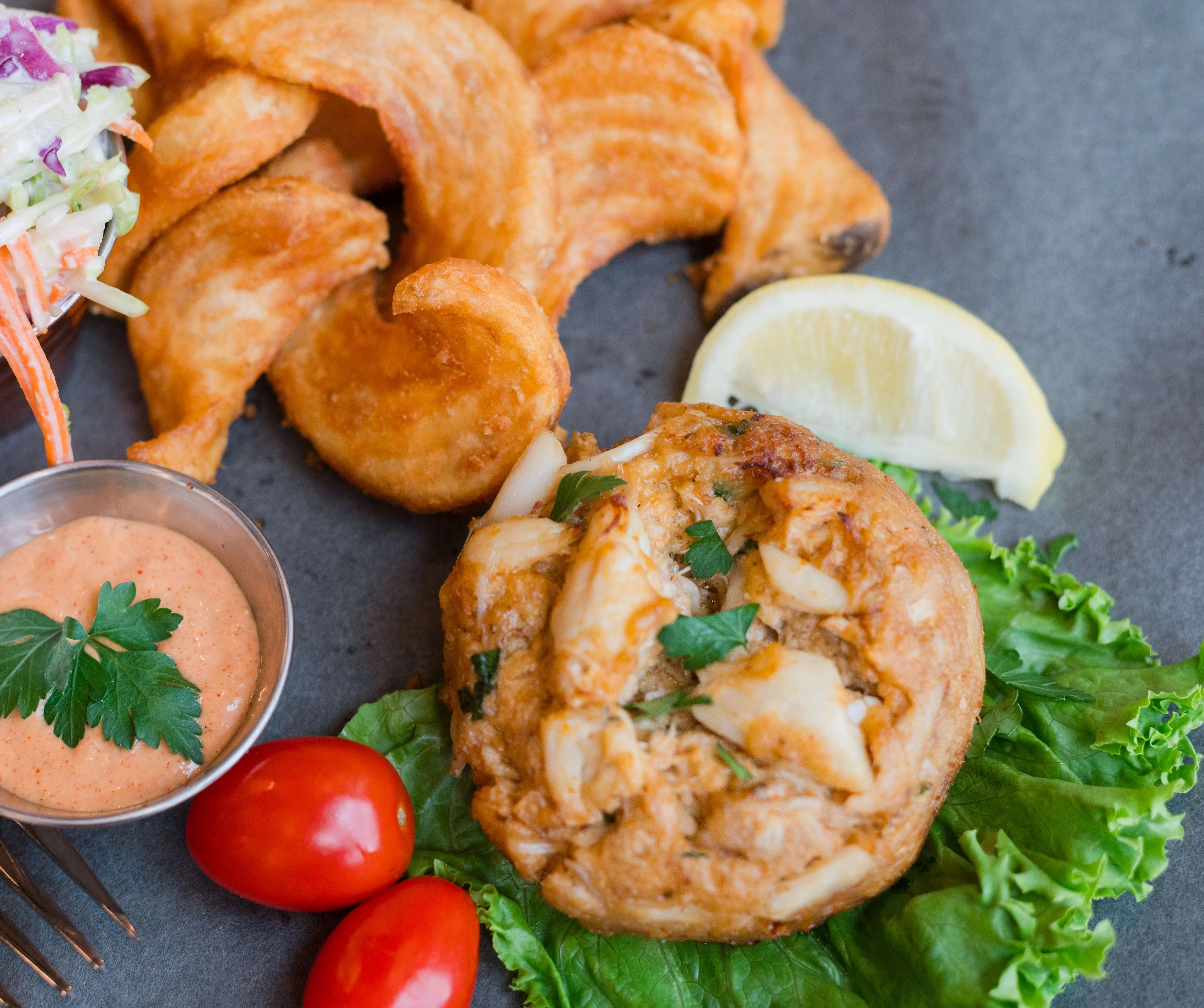 A plate featuring a crab cake on a bed of lettuce, accompanied by curly fries, a coleslaw side, a small bowl of sauce, cherry tomatoes, and a lemon wedge.