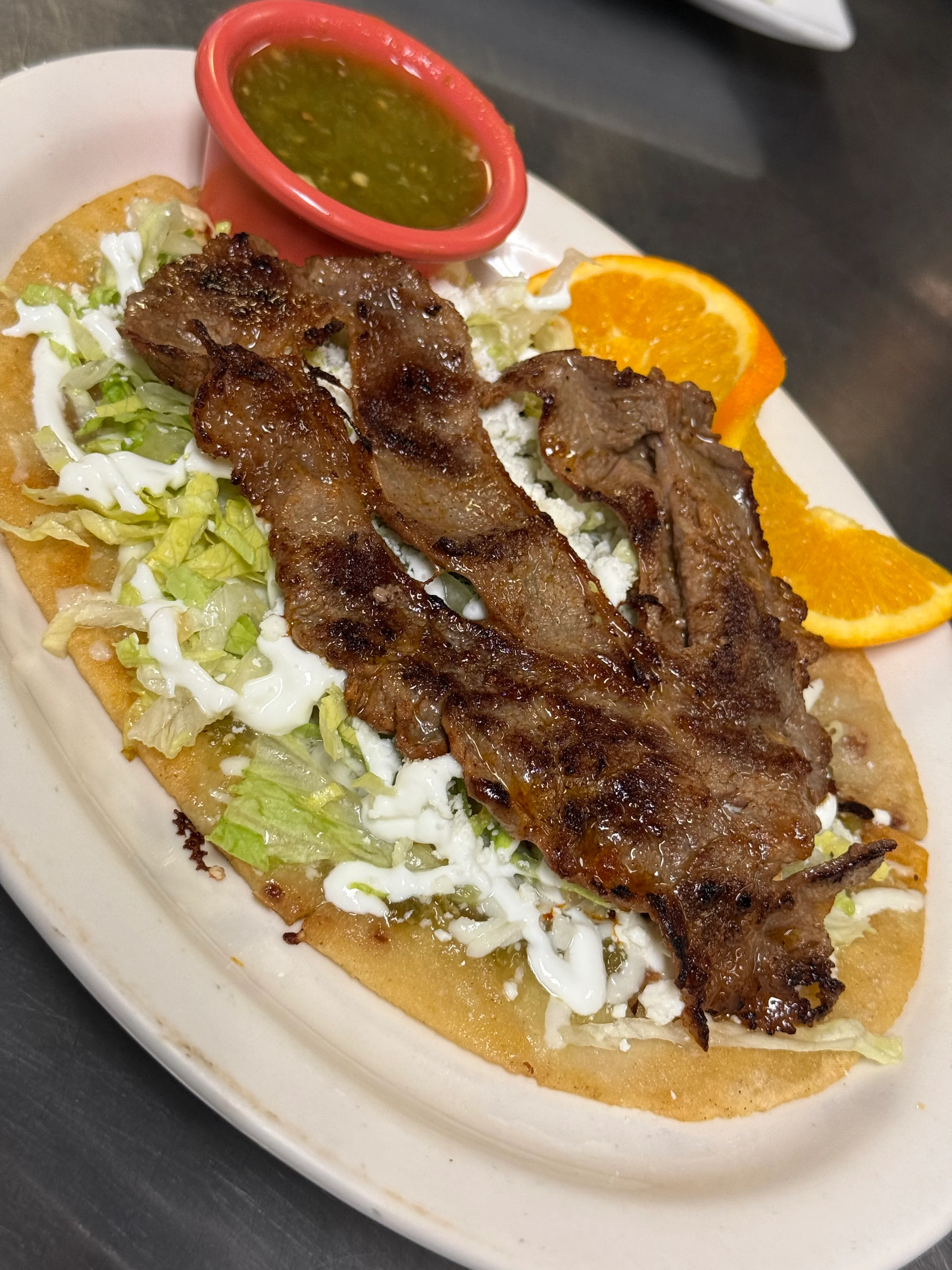A flatbread topped with grilled meat, lettuce, and cream, served with a small bowl of green sauce and orange slices.