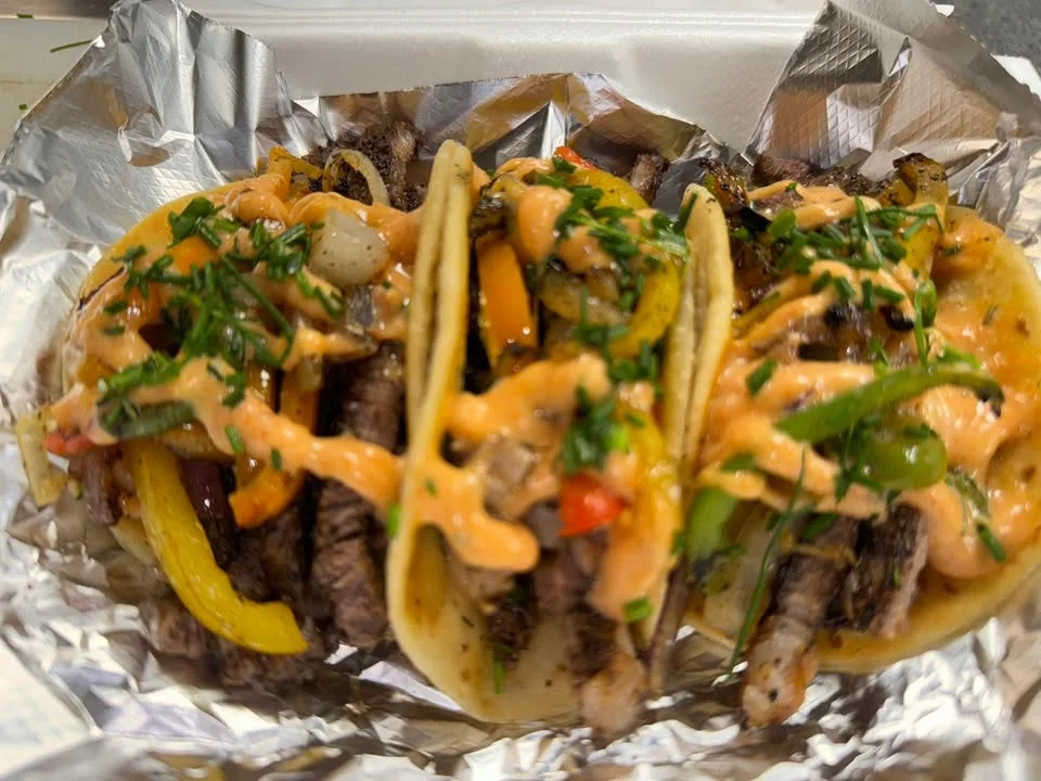 Three tacos filled with grilled beef, colorful bell peppers, and drizzled with a creamy sauce, garnished with herbs, served on a foil-lined tray.