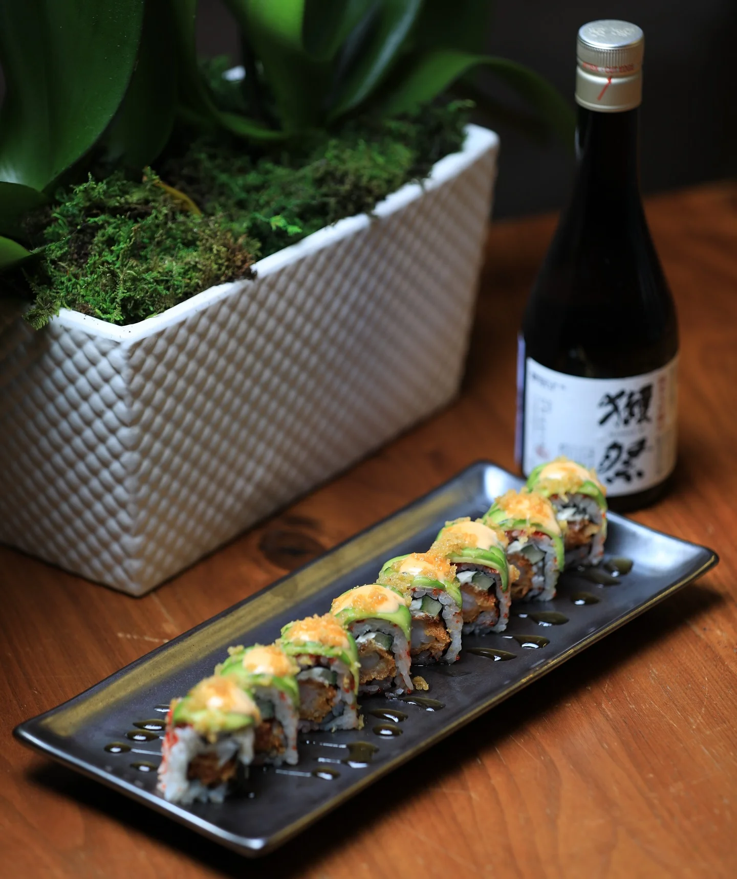 A row of specialty sushi rolls topped with avocado, spicy mayo, and crunchy tempura flakes, served on a black rectangular plate alongside a bottle of sake and a potted plant.