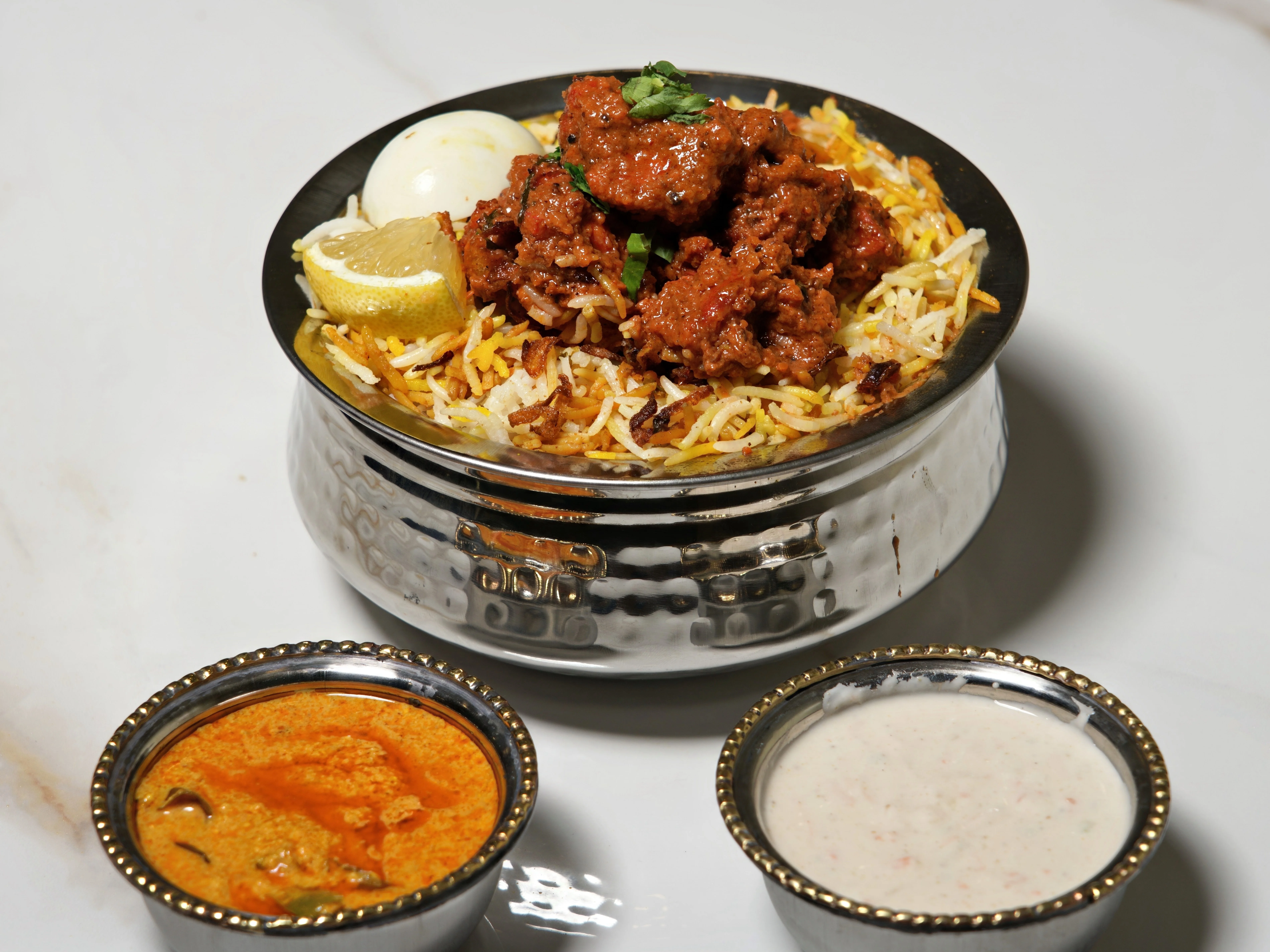 A bowl of biryani topped with tender meat, served alongside yogurt and a spiced curry, garnished with lemon and fresh herbs.