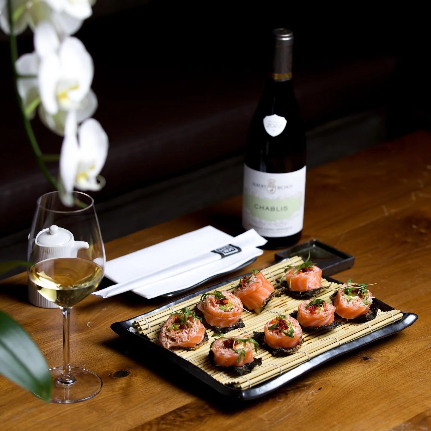 Salmon-wrapped sushi rolls presented on a bamboo mat, served on a wooden table alongside a bottle and glass of Chablis wine.