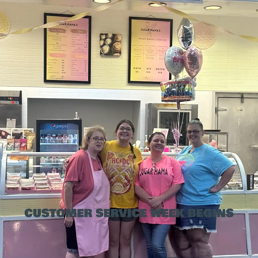 Four women smiling together inside Sugar Mama’s bakery, standing in front of the counter and menu boards. A birthday balloon bouquet is behind them, and the text “Customer Service Week Begins” is written across the photo.