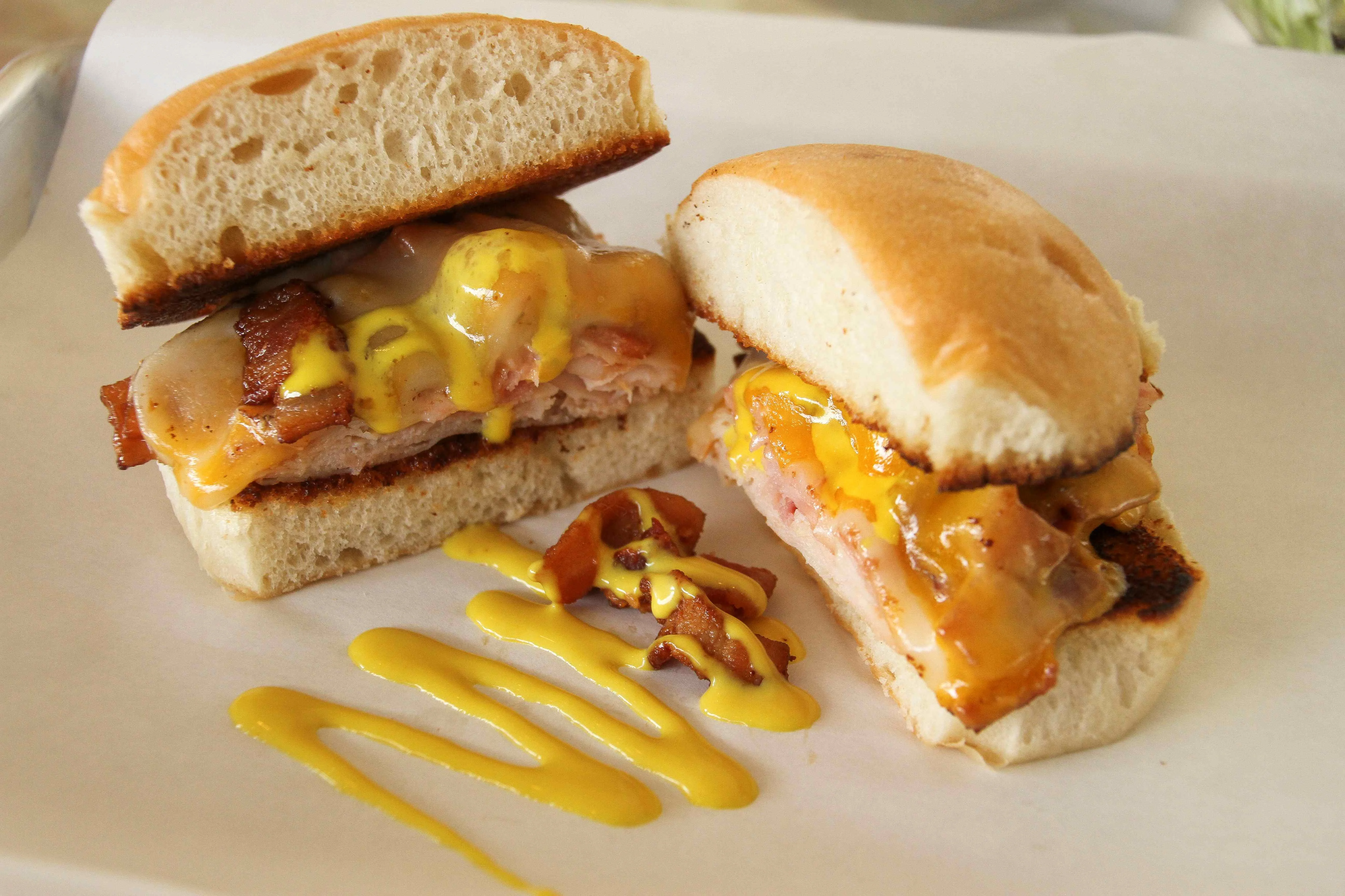 Toasted sandwich with sliced turkey, melted cheddar and Swiss cheese, crispy bacon, and mustard on a soft bun, served on parchment paper with a drizzle of mustard and bacon crumbles.