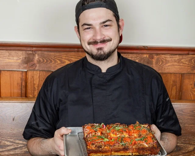 A sandwich made with a whole grain roll filled with fried chicken, marinara sauce, and melted cheese, served with a side of potato chips.