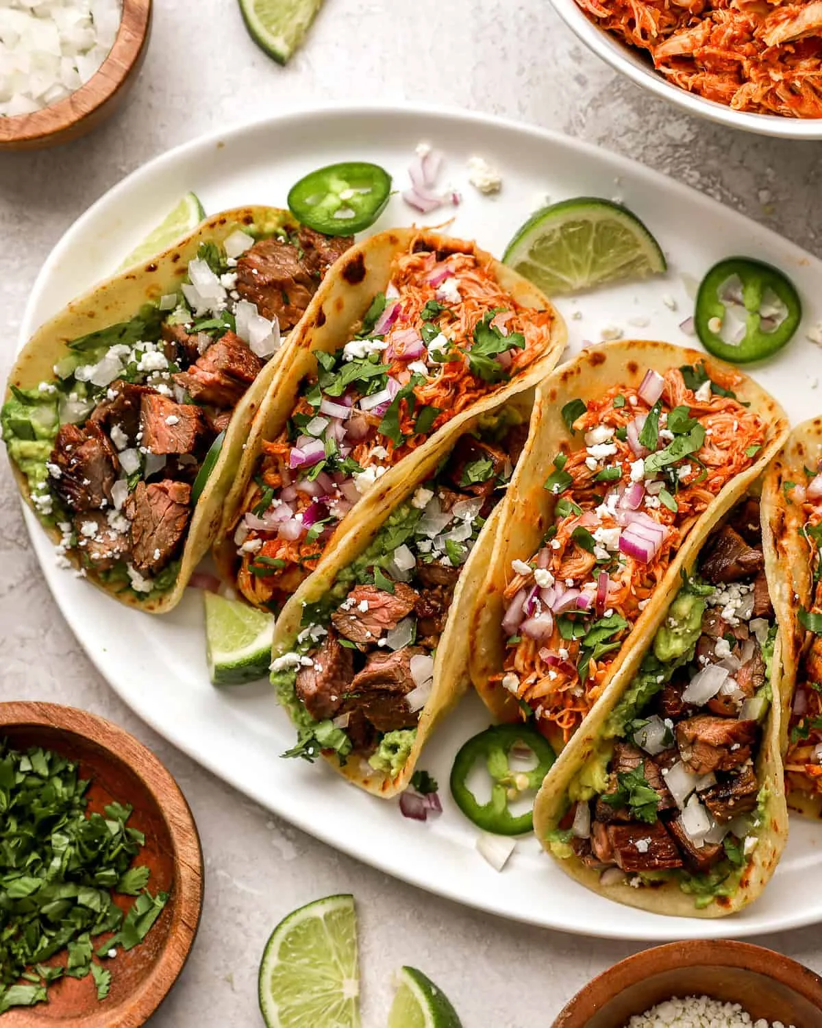 An assortment of tacos generously filled with various meats, garnished with onions, cilantro, and lime on a white platter.