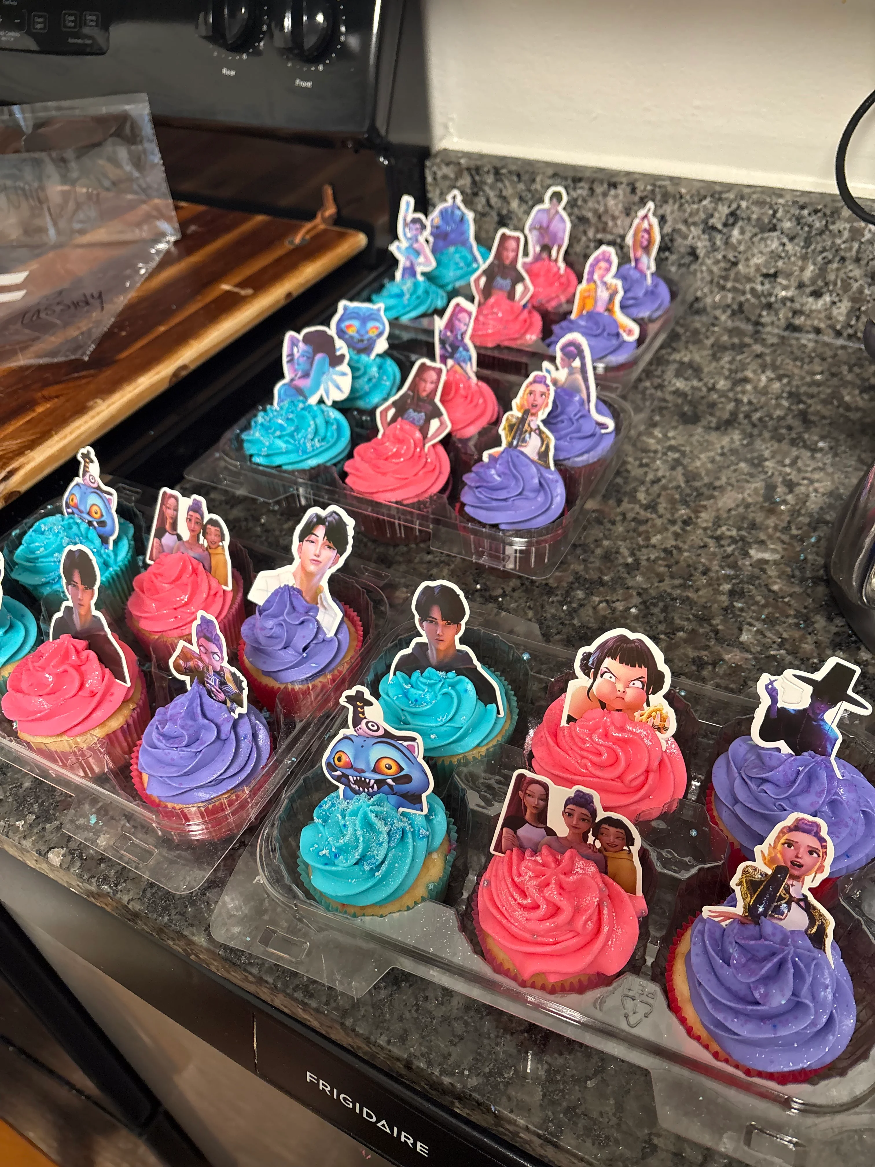 A variety of colorful cupcakes with blue and pink frosting topped with character picks, arranged on a kitchen counter.