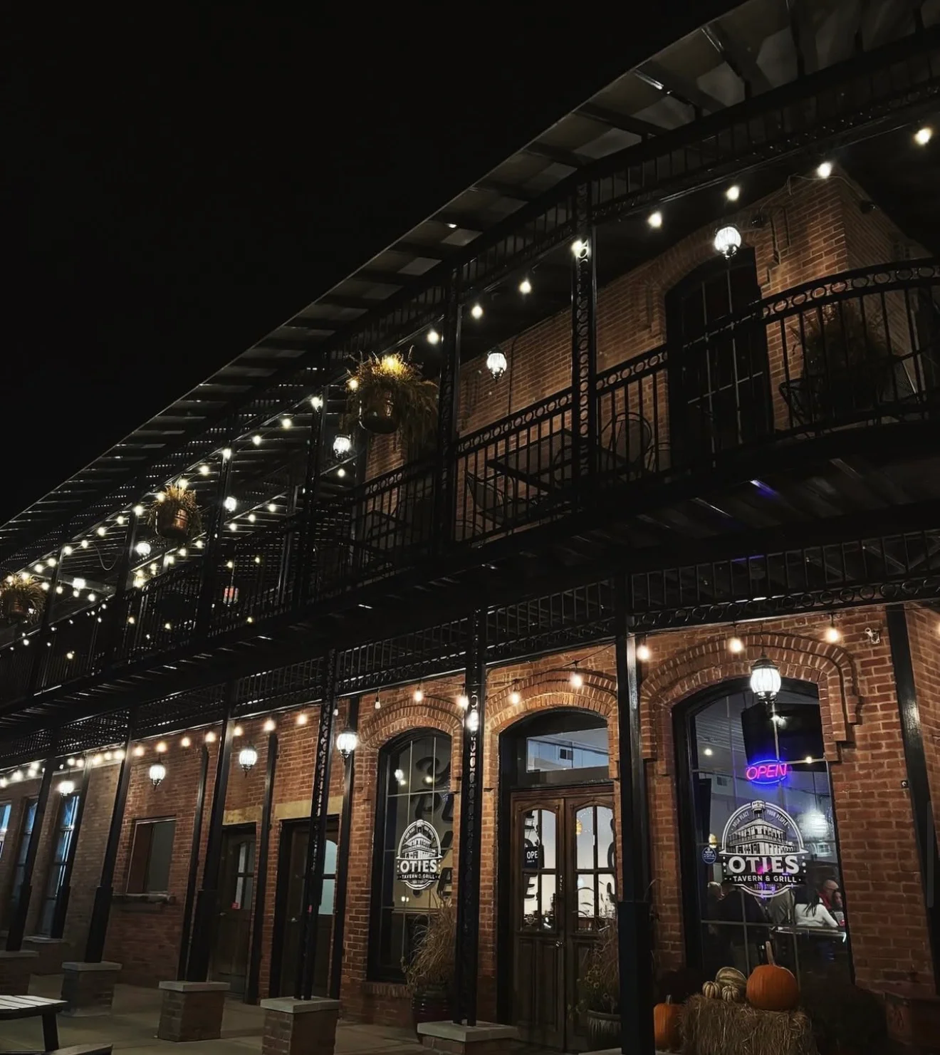 A rustic brick building illuminated by decorative string lights at night. Large windows and an 'Open' sign indicate a welcoming atmosphere.