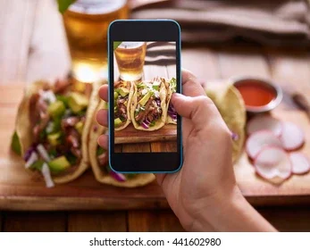Hand holding a smartphone capturing vibrant tacos on a wooden table