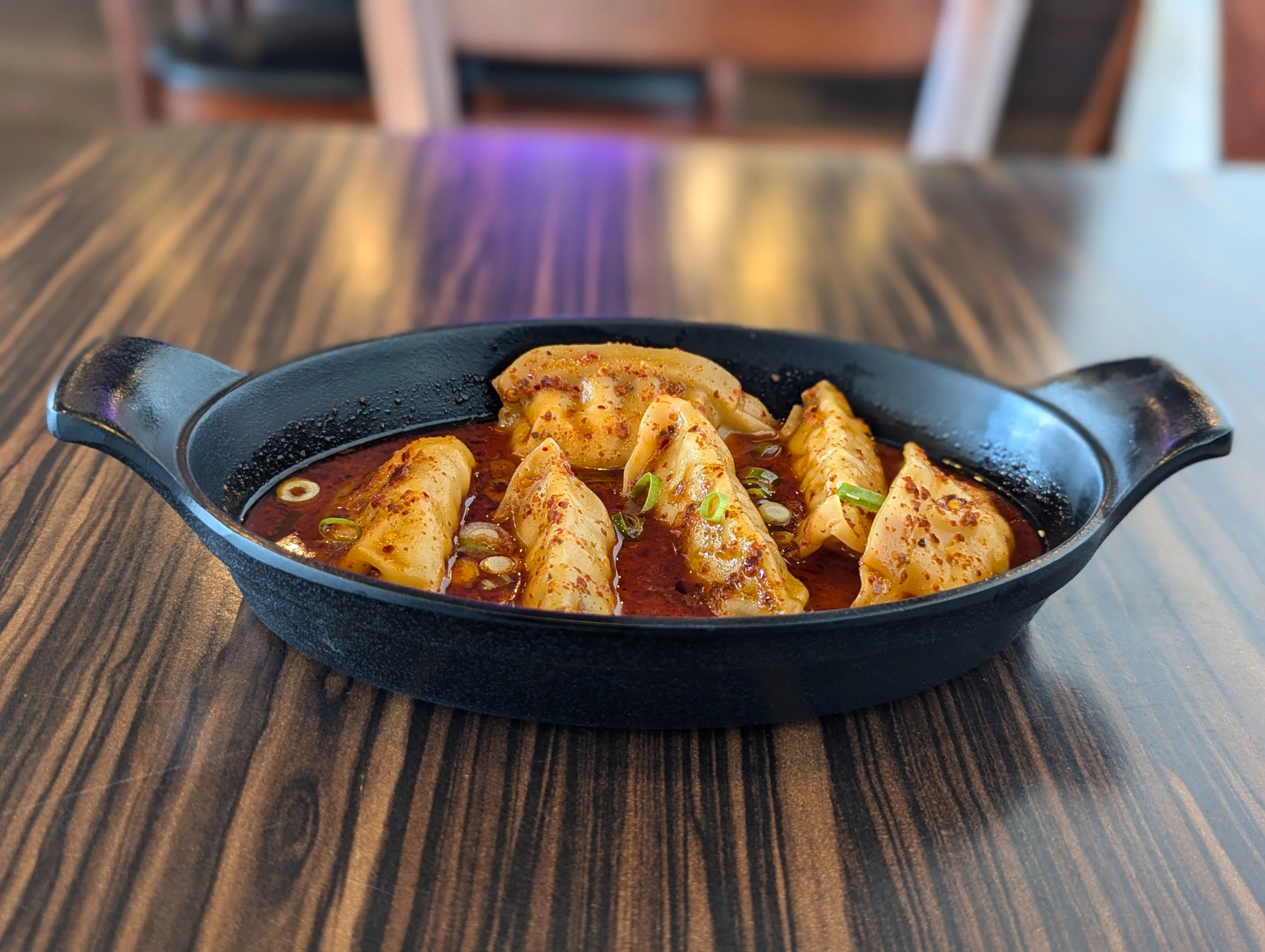 Spicy dumplings served in a black cast-iron dish at Bento Kitchen Cravings, topped with chili flakes and green onions in a rich, flavorful sauce, set on a wooden table, showcasing a bold and savory appetizer.