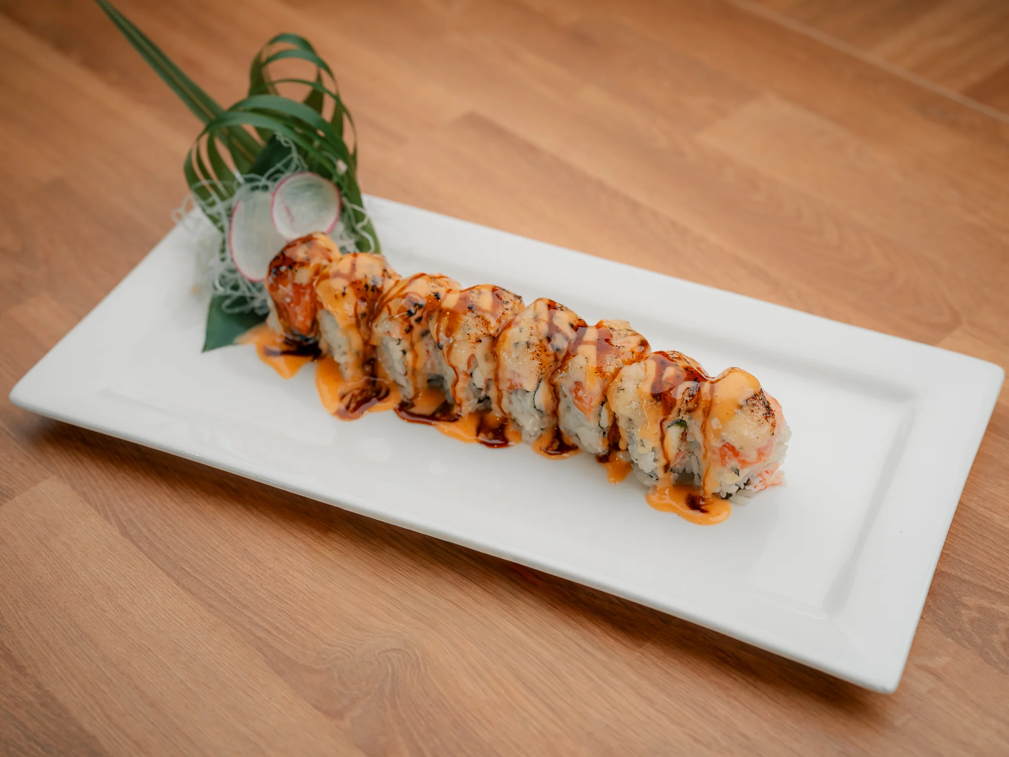 A beautifully plated sushi roll topped with a drizzle of sauce and garnished with greens and radish slices.