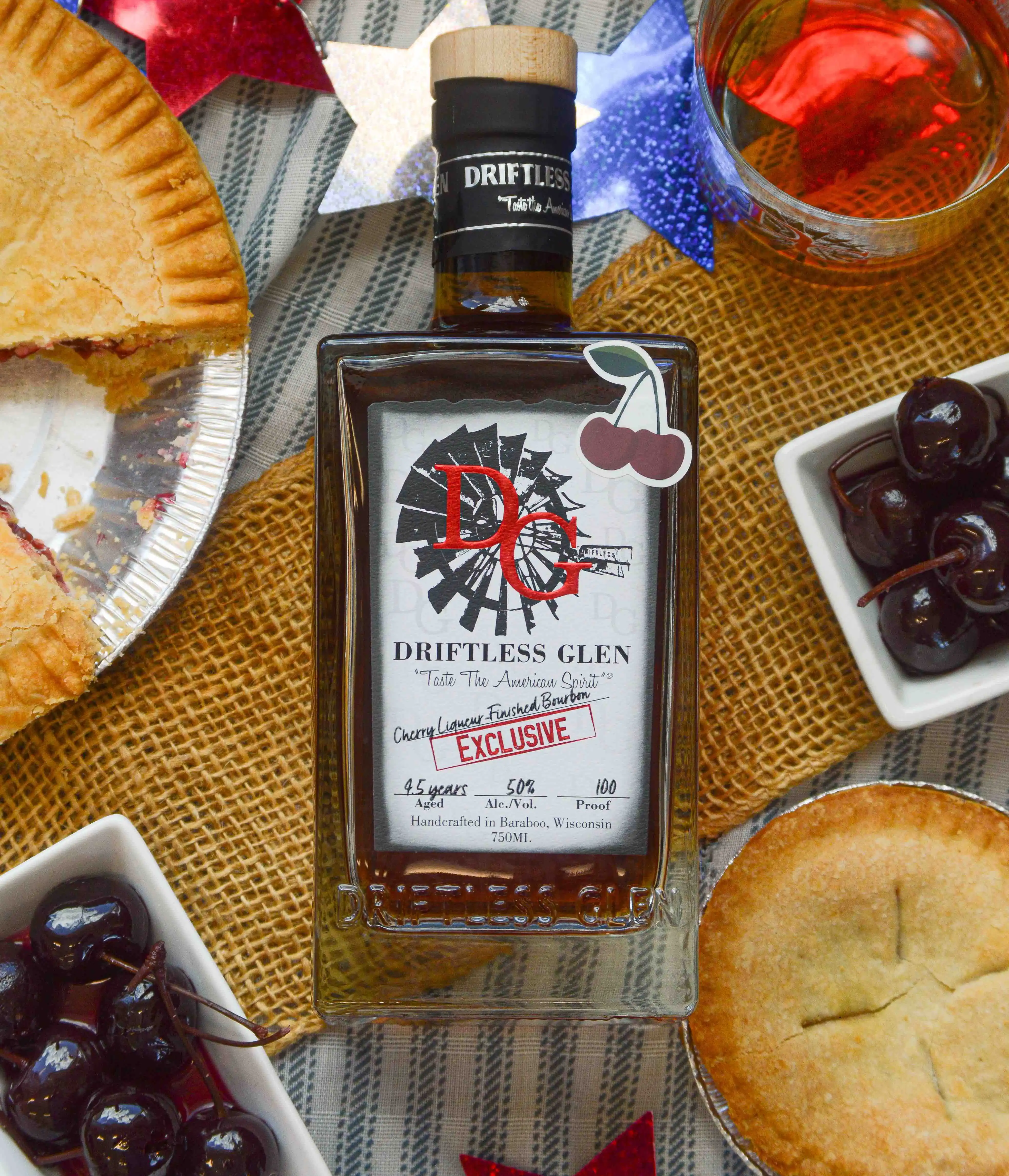 A bottle of Cherry Liqueur-Finished Bourbon sits next to a cherry pie and cherries.