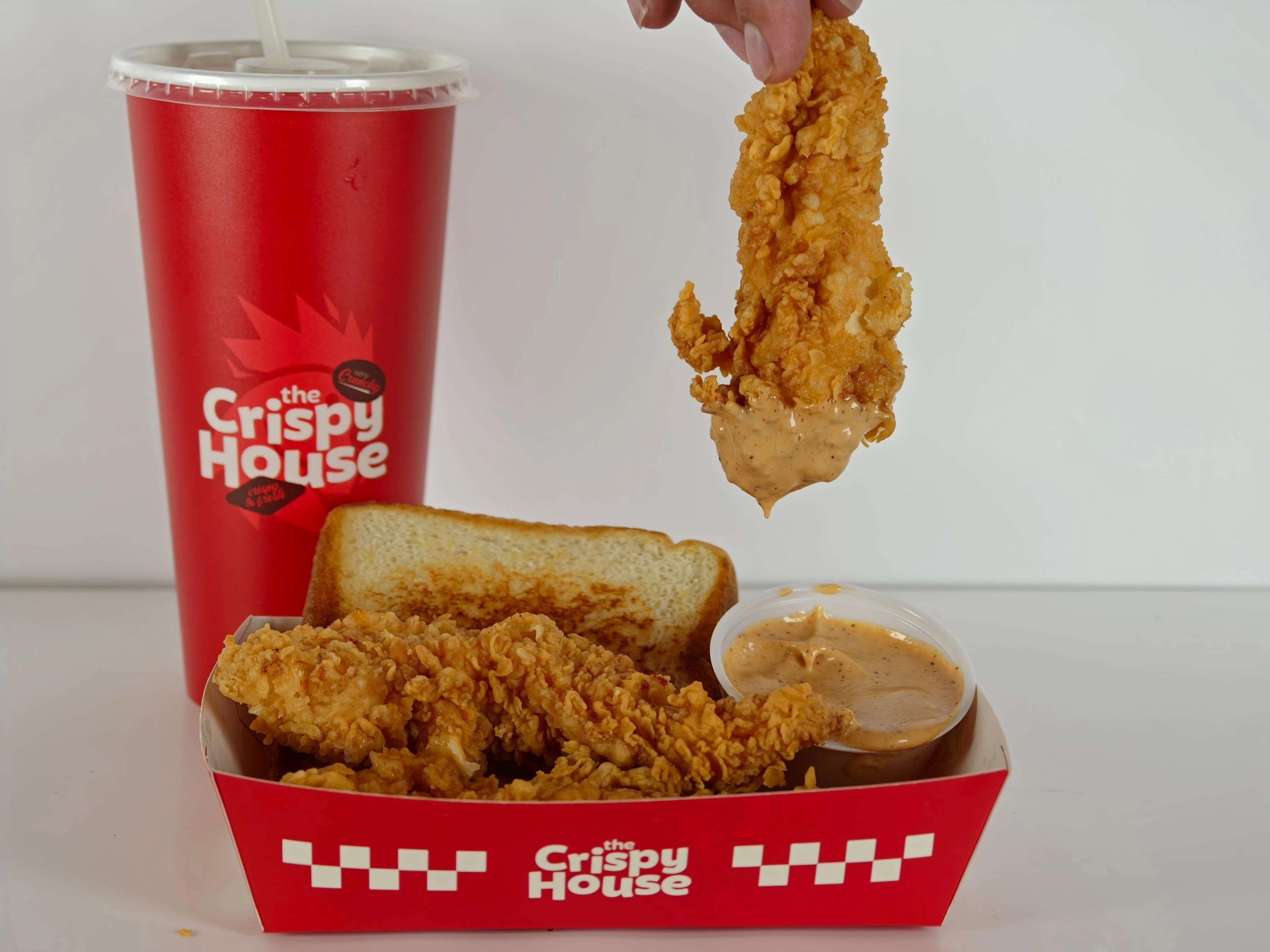 A serving of crispy chicken tenders being dipped into a sauce, accompanied by a slice of bread and a drink.