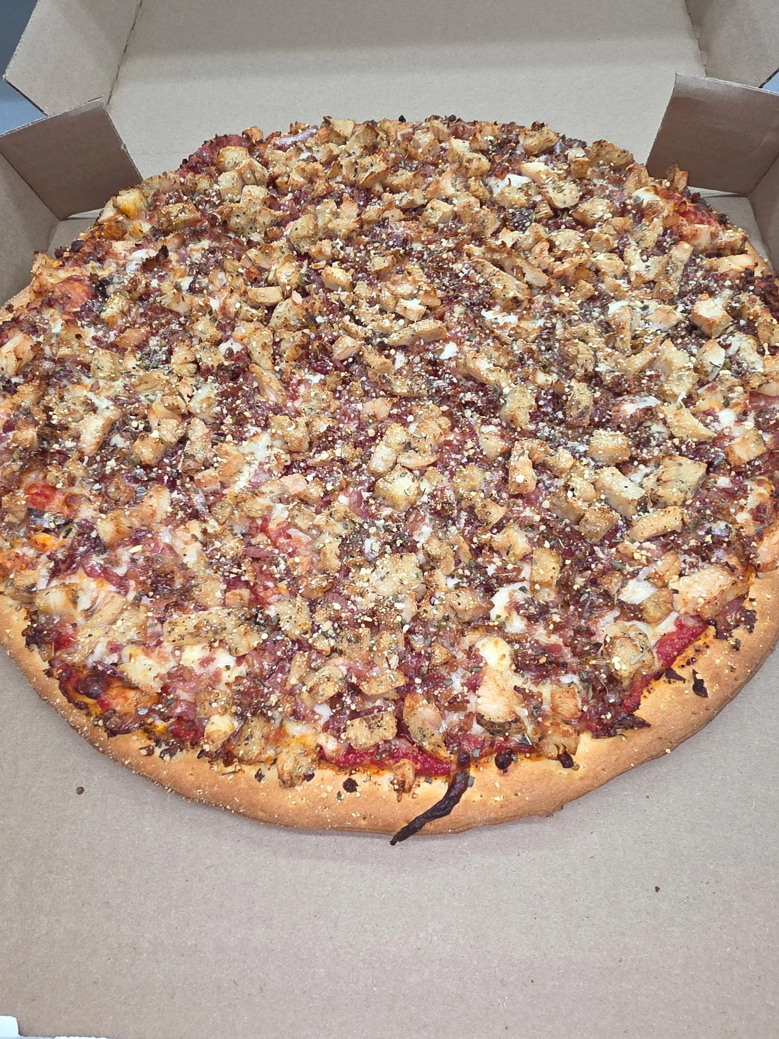 A pizza in a cardboard box, topped with chopped grilled chicken and seasoning.