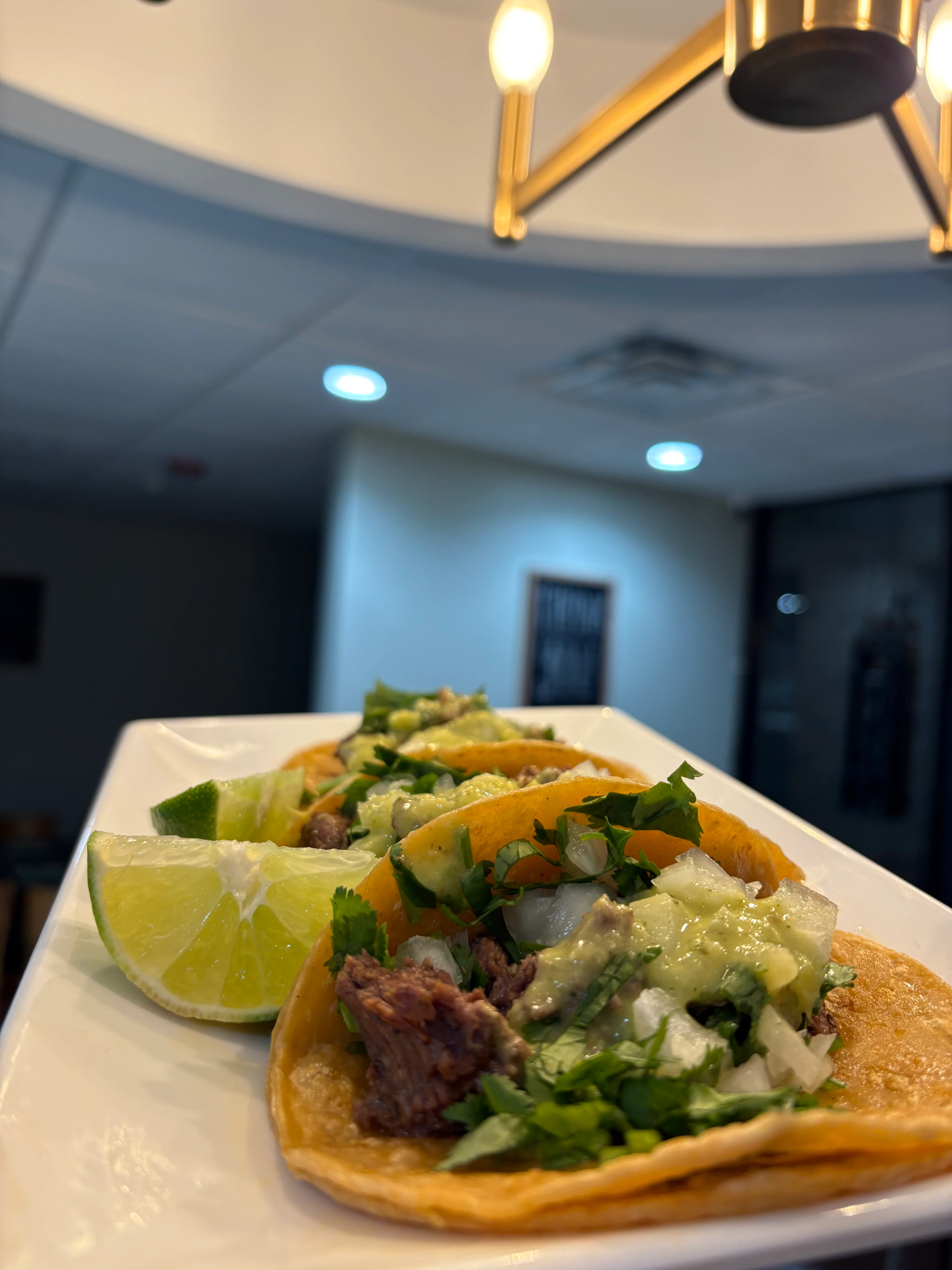 Three tacos filled with seasoned beef and topped with onions, cilantro, and a drizzle of sauce, accompanied by lime wedges.