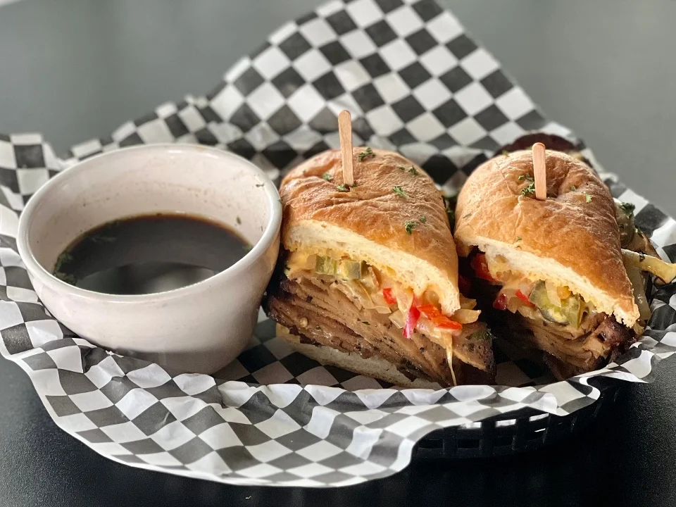 A plate with two sandwiches garnished with toothpicks, served with a small bowl of dipping sauce on a checkered paper.