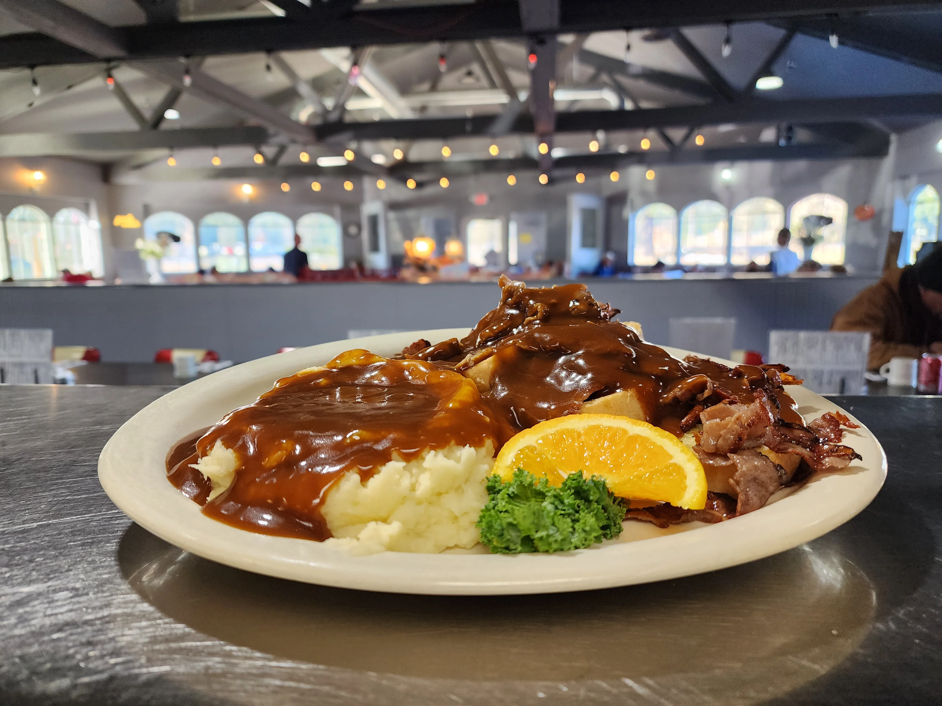 A plate of mashed potatoes topped with savory gravy and served with sliced meat, garnished with an orange slice and kale, set in a dining area.