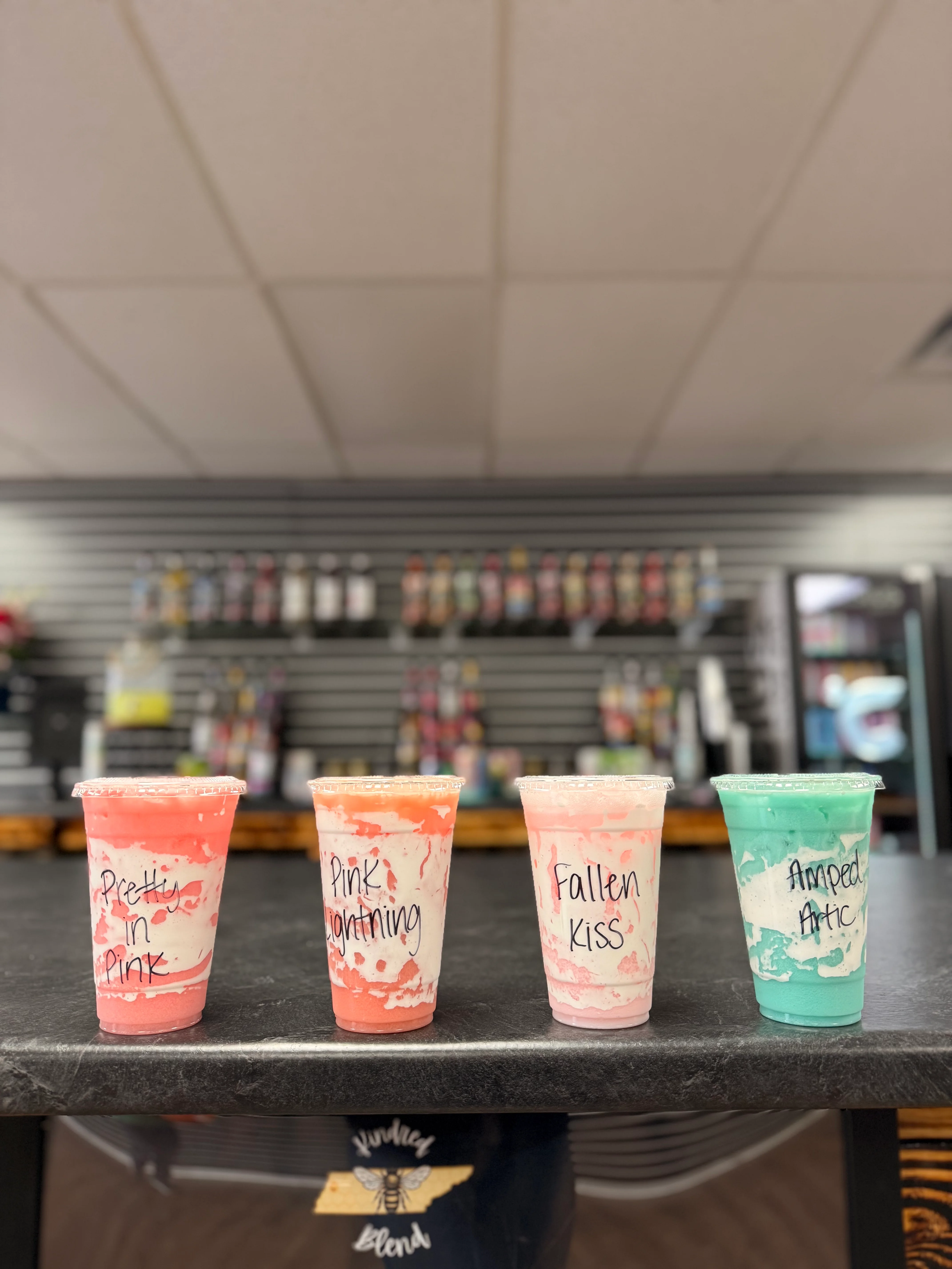 Four colorful drinks in clear cups, labeled with names, arranged neatly on a countertop with various flavored syrups in the background.
