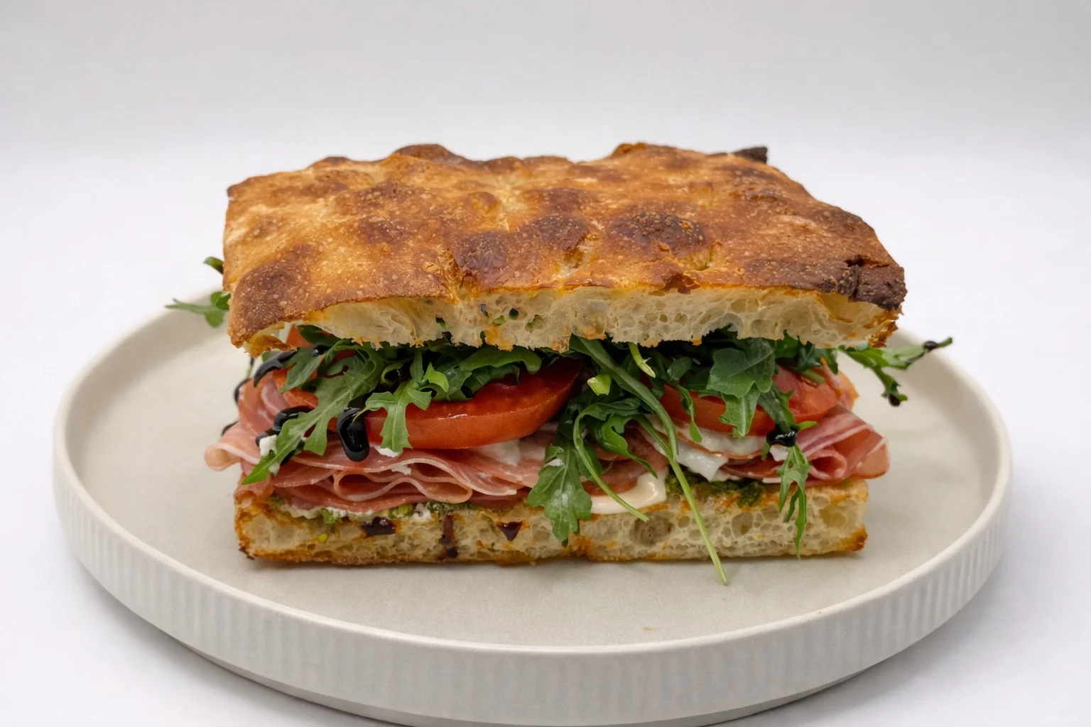 A colorful sandwich layered with cold cuts, fresh tomatoes, and arugula on a toasted bread base, displayed on a beige plate.