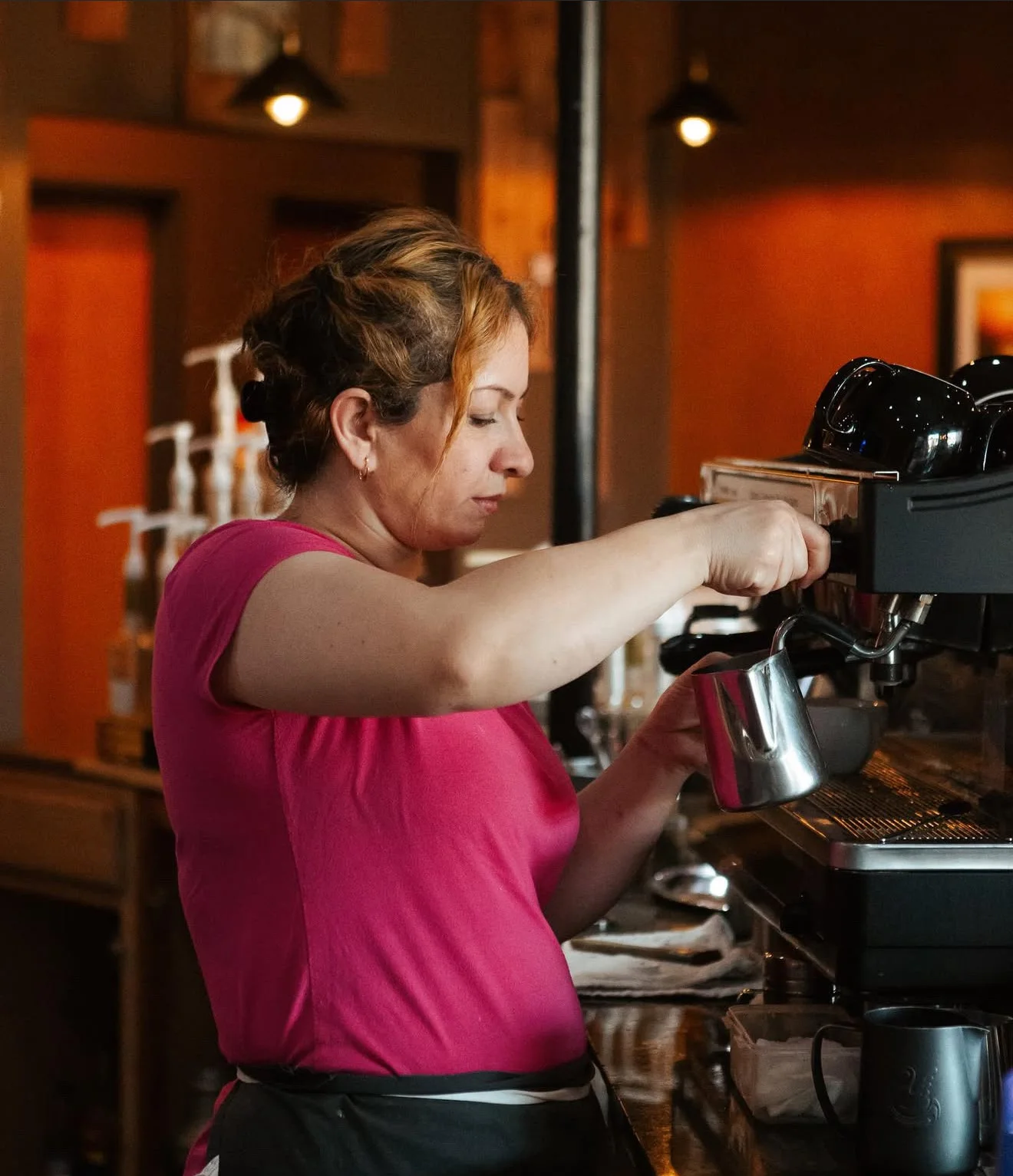 A barista pouring steamed milk into an espresso machine, focused on crafting a coffee beverage in a cozy café environment.