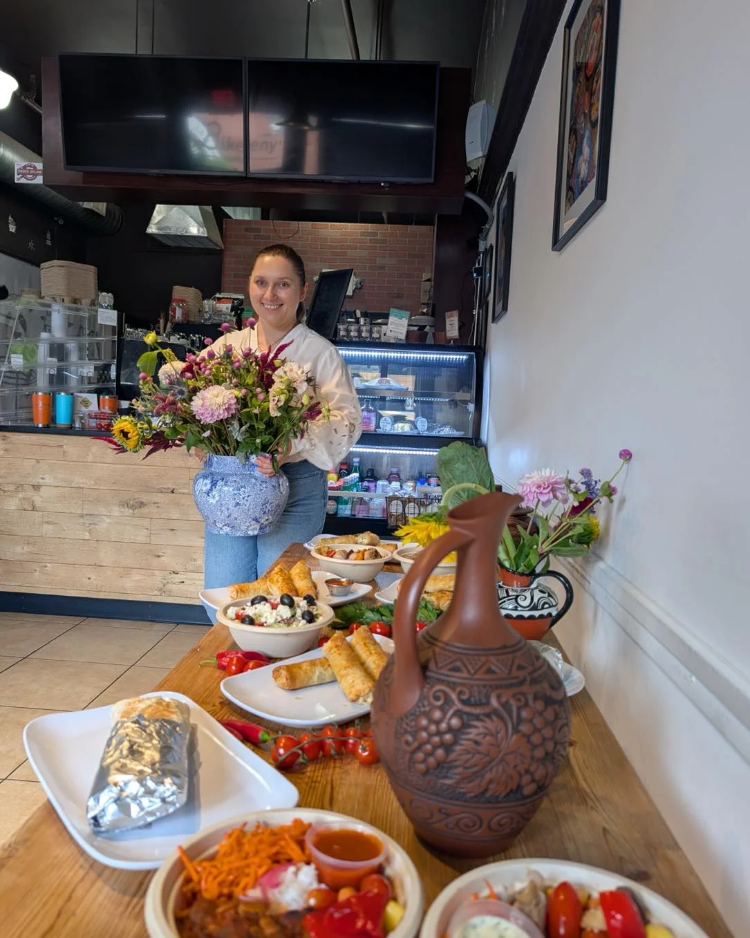A display of various dishes on a wooden table, with a woman holding a vase of flowers. The scene includes colorful food items and a decorative ceramic jug.