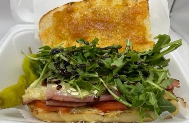 A sandwich filled with layers of ham, cheese, fresh greens, and tomatoes, with a toasted bun and pickles on the side.