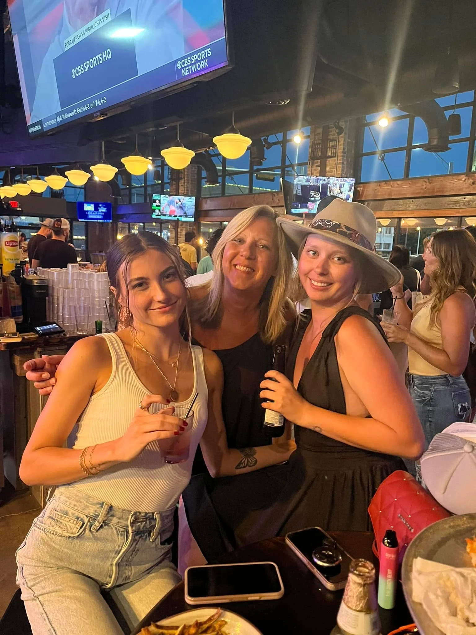 Three friends enjoying drinks at a lively bar with sports on TV