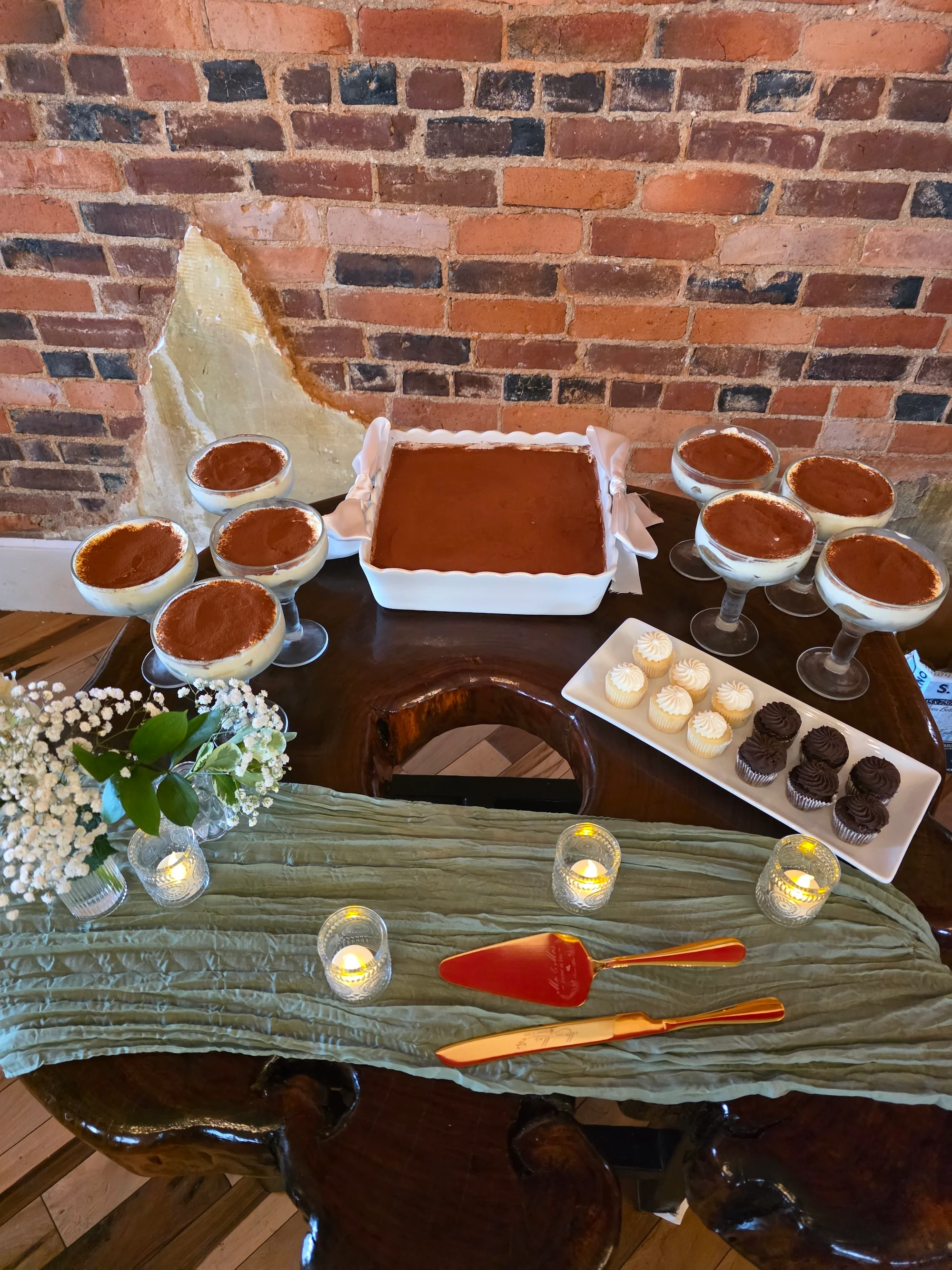 A dessert spread featuring several servings of tiramisu in cups alongside a larger dish of tiramisu. The table is decorated with flowers and lit candles, creating a cozy atmosphere.