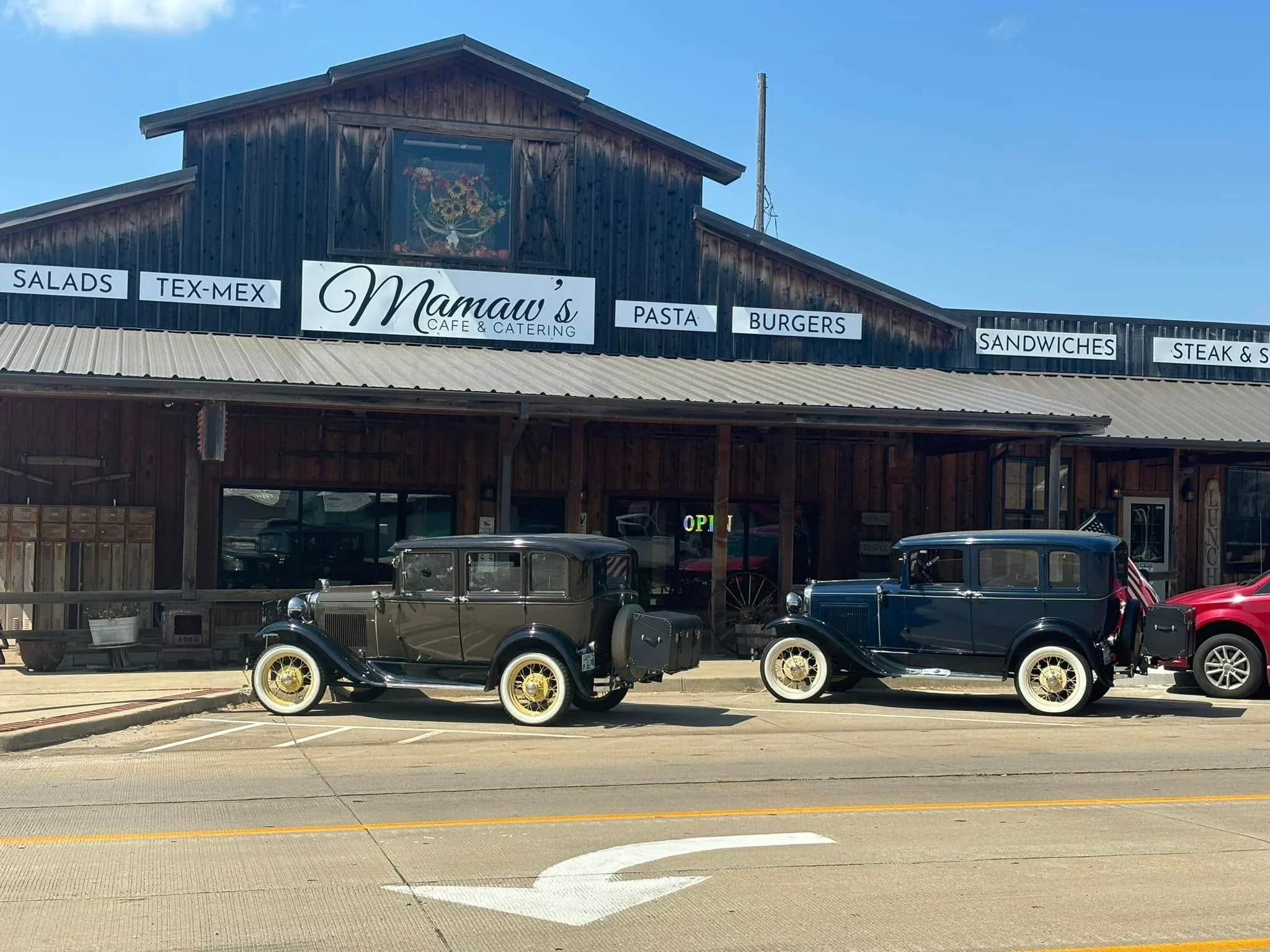 A vintage café and catering establishment with two classic cars parked in front. The building features rustic wooden architecture and signage indicating various food offerings.