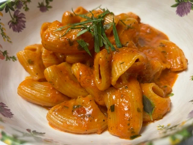 A bowl of pasta coated in a rich tomato sauce and garnished with herbs. The noodles are tubular and the dish is colorful.