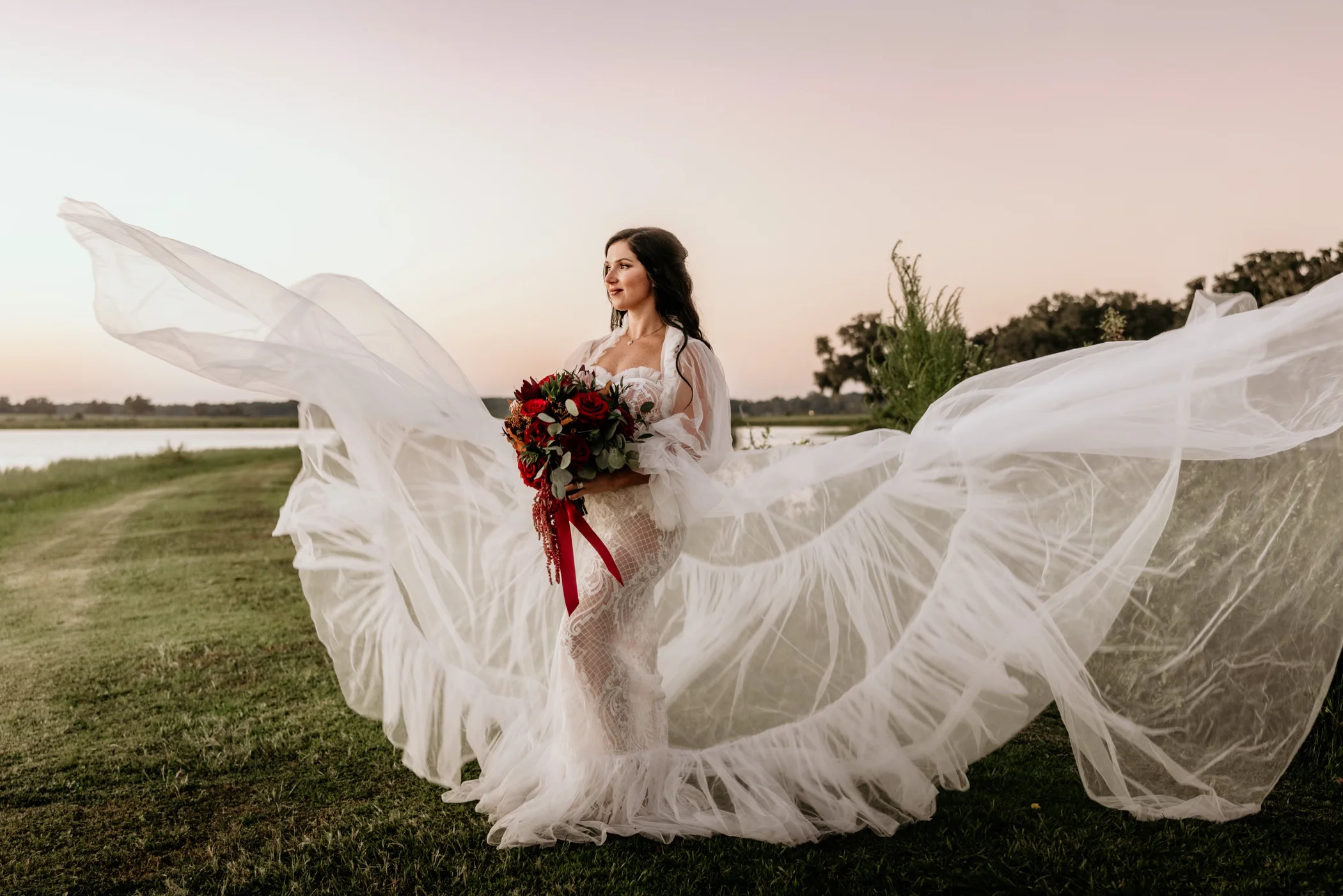 Bride in flowing gown holding bouquet by the water at sunset