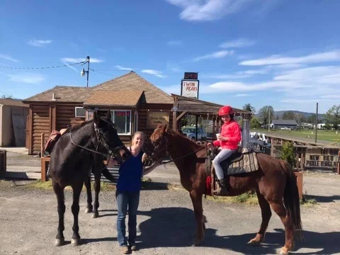 Two horses with riders outside a rustic restaurant on a sunny day