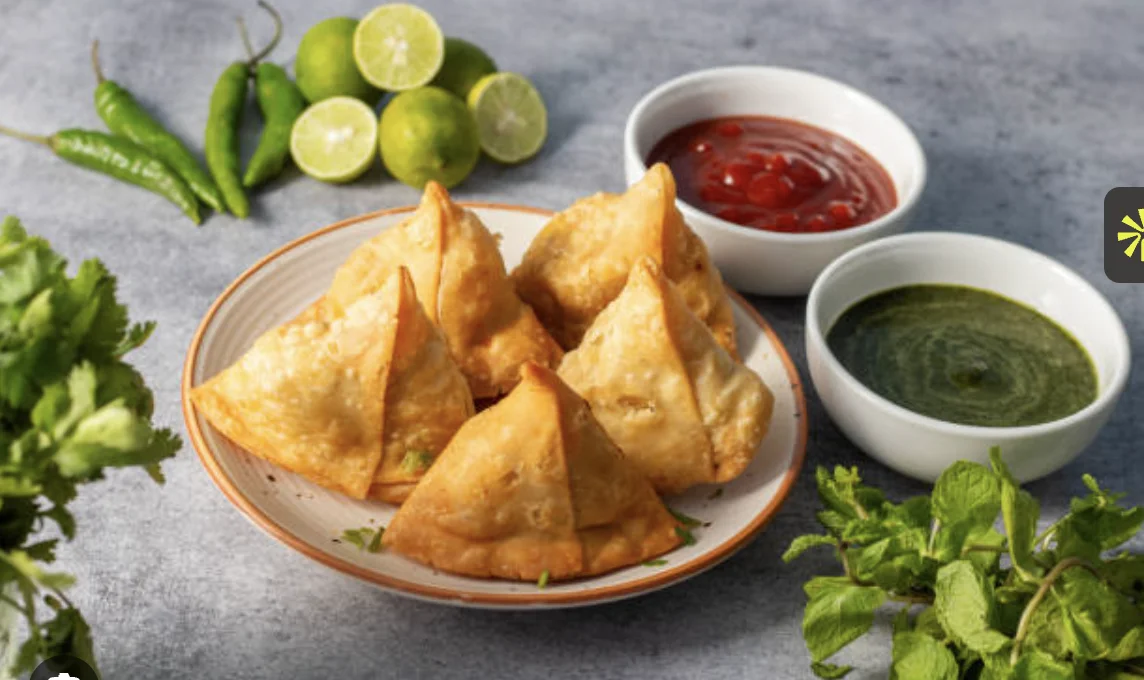 A serving of golden-brown samosas arranged on a plate, accompanied by green and red chutneys, and garnished with fresh lime and green chilies.
