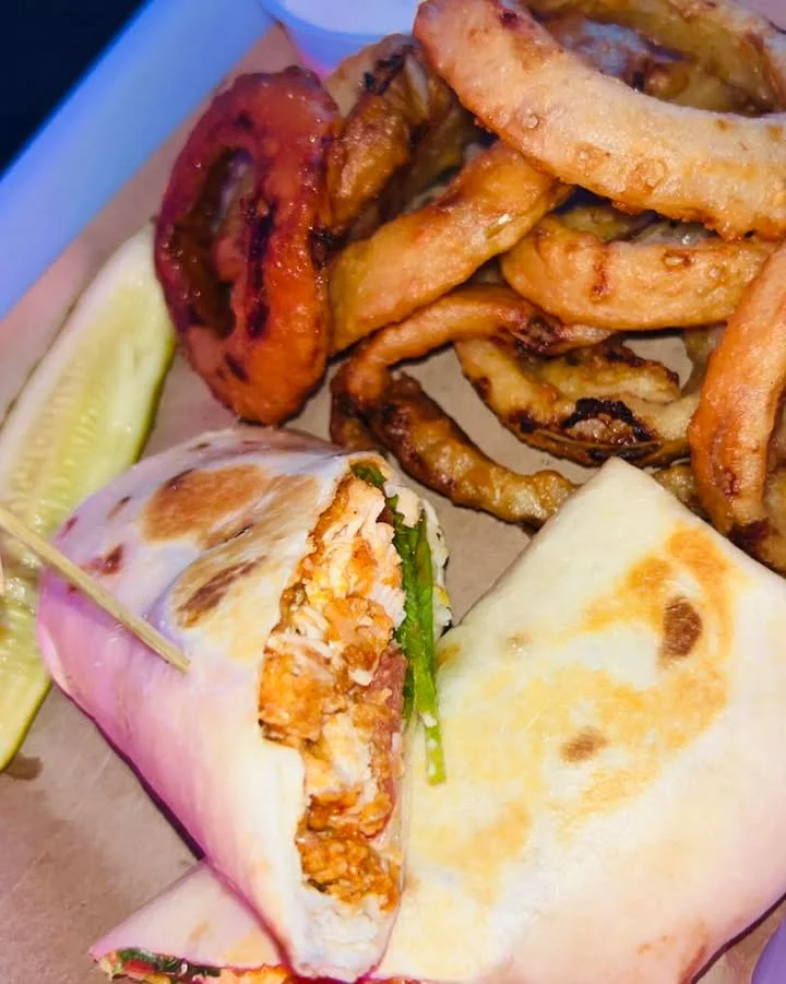 A serving of wraps filled with a mixture of ingredients, accompanied by crispy onion rings and a slice of pickle.