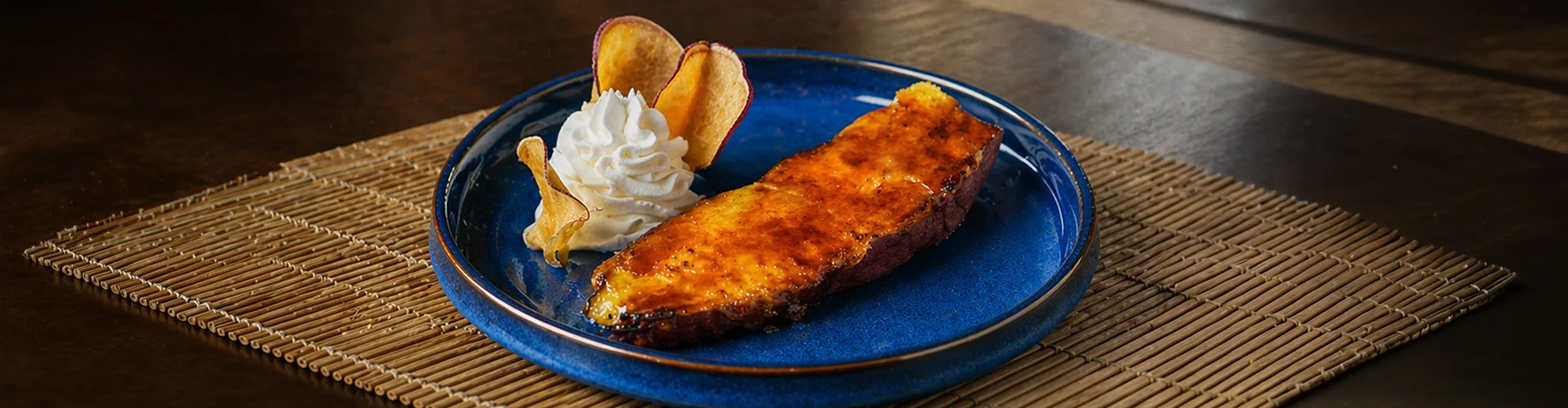 Two halves of a roasted sweet potato with a caramelized, golden-brown topping and a scoop of vanilla ice cream on a bold blue plate. There is also a text overlay which leads to the desserts menu.