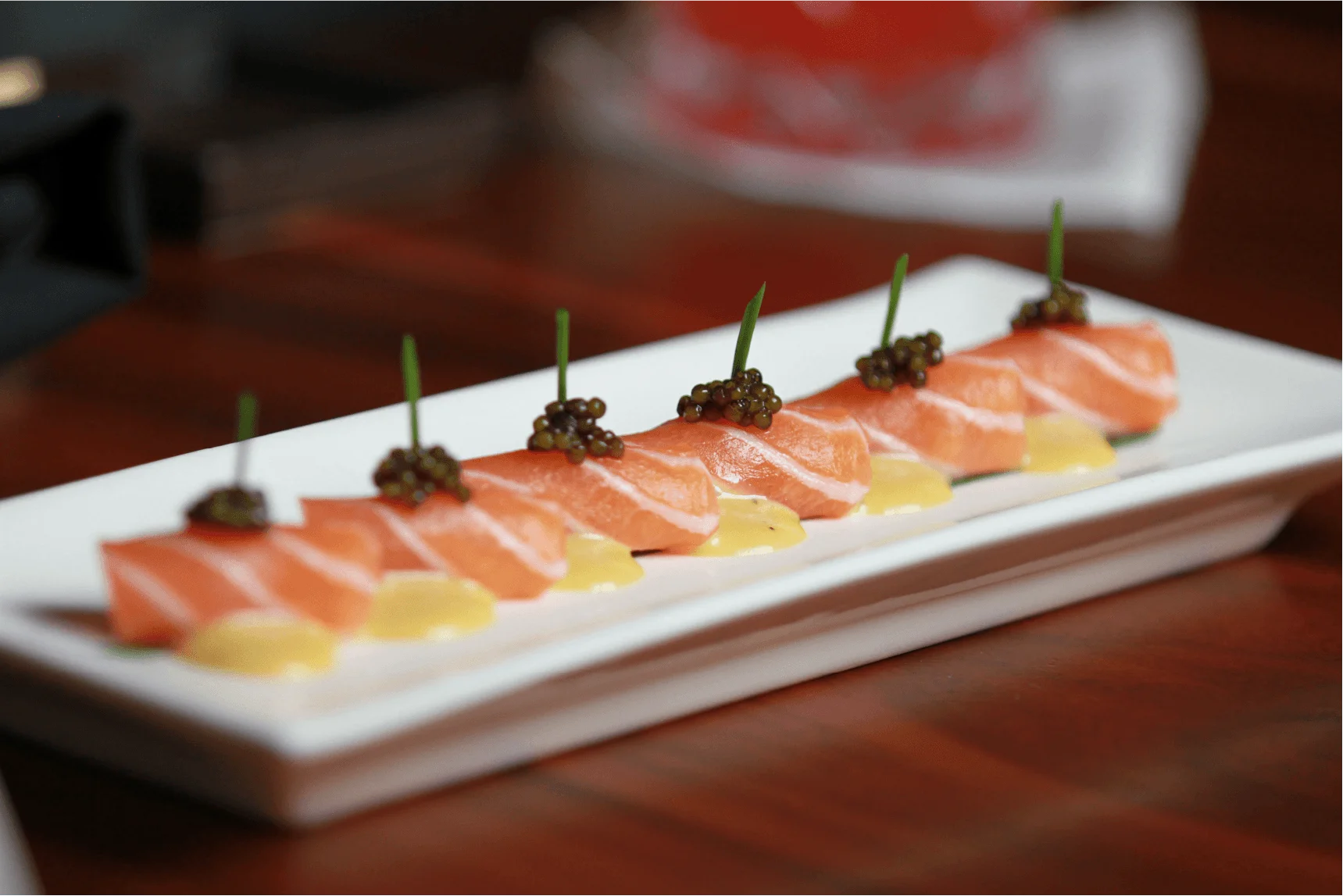 A row of six delicate salmon sashimi pieces arranged neatly on a long white rectangular plate, each piece resting on a small dollop of yellow sauce and topped with a cluster of dark caviar and a single green chive sprout.