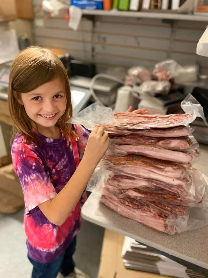 A child smiling while posing with a stack of packaged meats on a table.