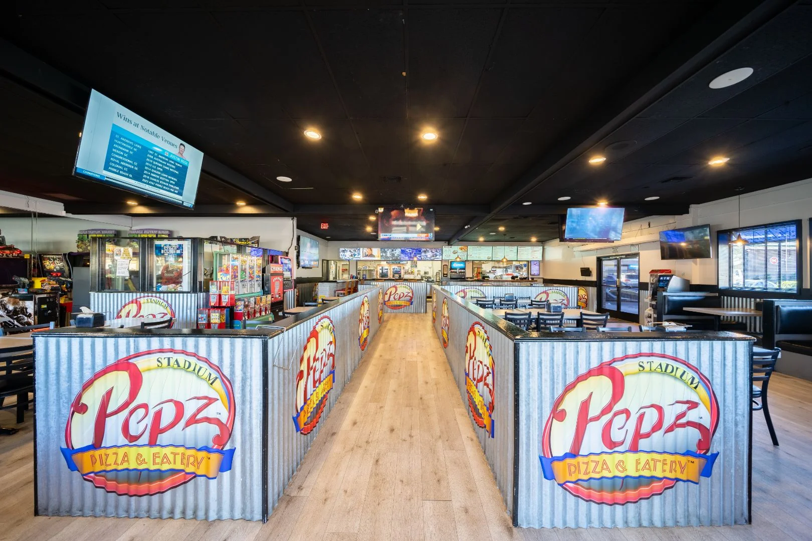 Interior of a casual pizza restaurant with a central walkway, corrugated metal partitions featuring logo signage, arcade games to the side, and seating areas with TVs mounted overhead.