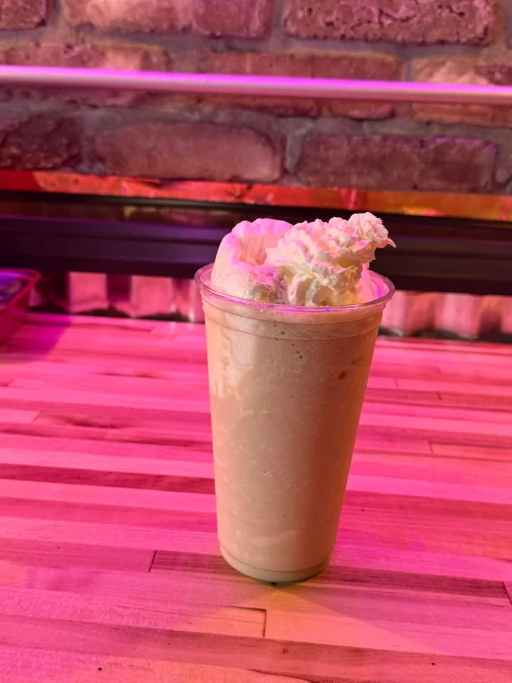 A creamy beverage in a clear cup topped with whipped cream, sitting on a wooden countertop with a textured background.