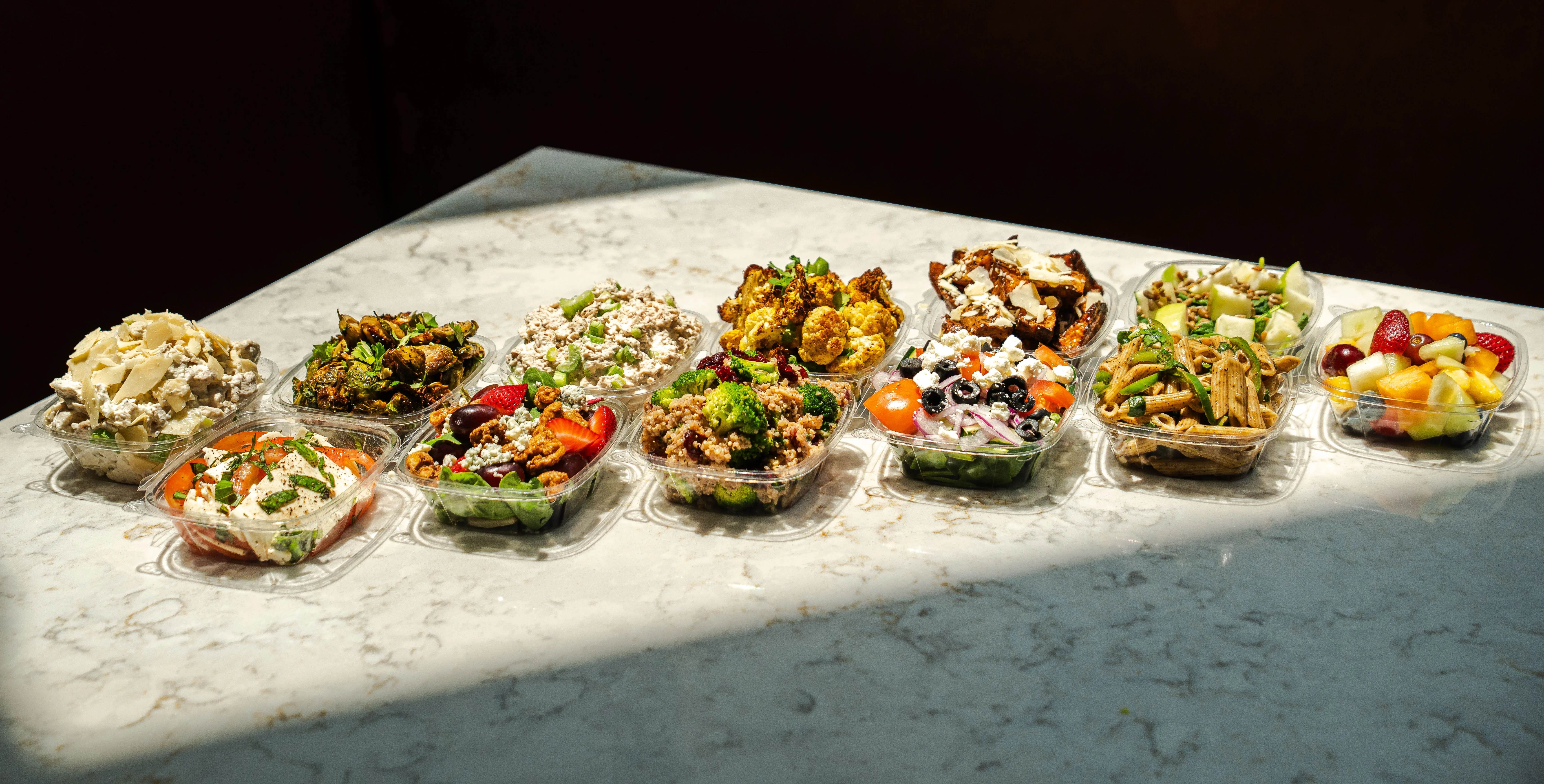 Devon & Blakely's colorful spread of prepared salads, roasted vegetables, pasta dishes, and fresh fruit in clear takeout containers on a marble counter.