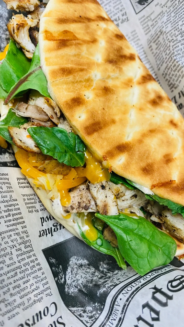 A sandwich made with grilled chicken, spinach, and cheese, served in a toasted flatbread, wrapped in newspaper for presentation.